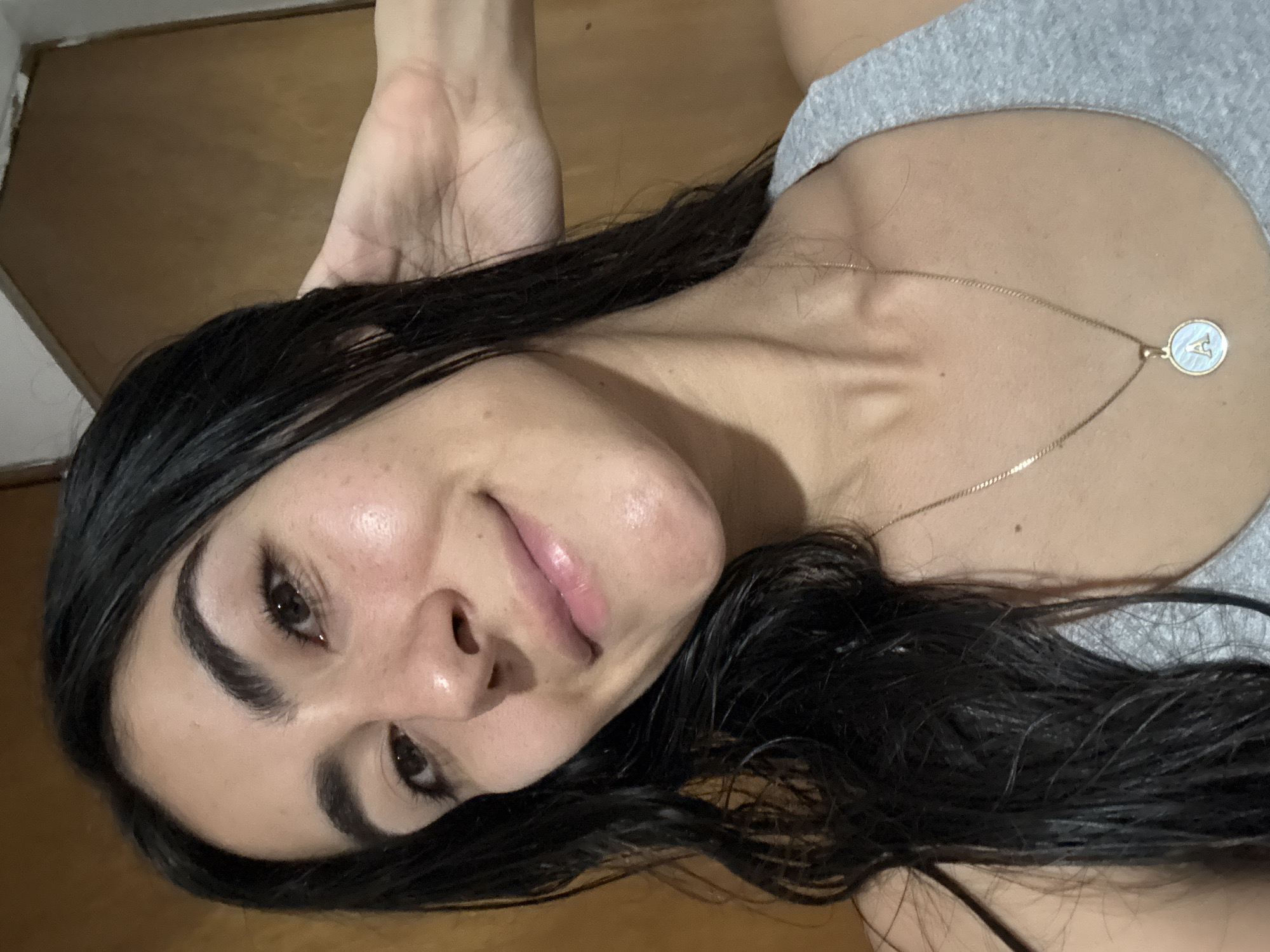 The image is a tilted, close-up portrait of a young woman, captured from the chest up. She appears to be lying down or lounging, with her head turned slightly towards the camera. Her long, dark hair, which looks somewhat damp or shiny, frames her face and rests on her shoulders. She wears a simple grey top, with a strap visible over her right shoulder. Around her neck is a delicate gold chain necklace, from which hangs a small, circular pendant with a white or light blue background, bordered in gold. A stylized capital letter 'A' is clearly visible on the pendant.
The woman has fair skin, with a natural, subtle sheen evident on her forehead, nose, and chin, suggesting a fresh or moisturized complexion. Her dark eyes are open and directed towards the viewer, complemented by well-groomed, dark eyebrows. Her lips are slightly parted in a gentle, warm smile, revealing a natural pink tone. Part of her left hand is visible near the upper left corner of the frame. The background is simple, featuring a wooden surface, possibly a floor or wall, and a glimpse of a light-colored wall or trim, indicating an indoor setting. The overall expression on her face is calm and content, suggesting a relaxed moment, likely in a casual home environment in Puebla, Mexico.