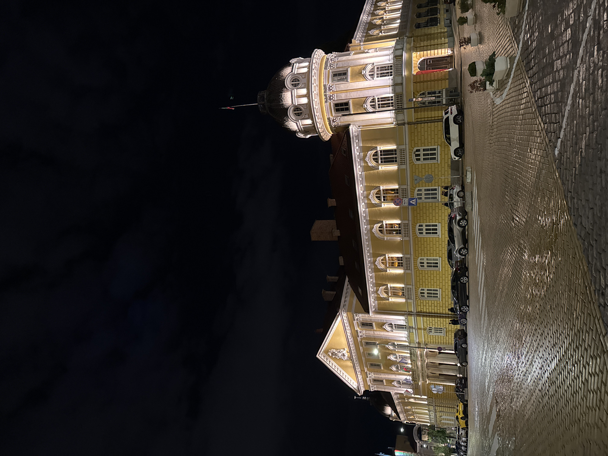 At night in Sofia, Bulgaria, a yellow building with a dome is lit up against a dark sky. The building has multiple windows and is adorned with white architectural details. Cars are parked along the cobblestone street in front of the building. The street is wet, reflecting the lights.