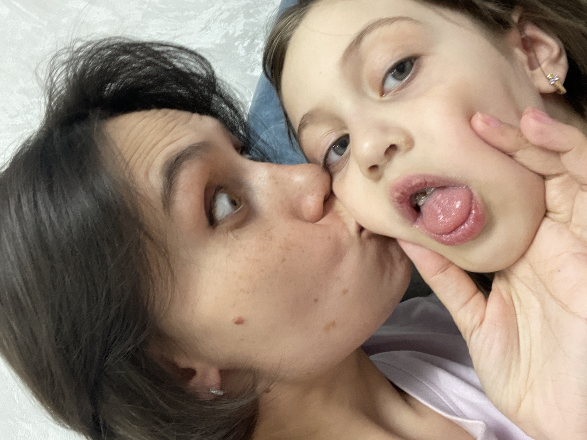 A close-up shot captures a light-skinned woman with dark hair and a fair-skinned child with light brown hair. The woman appears to be kissing the child's cheek while holding the child's face with one hand. The child is sticking their tongue out.
There is no information in the image that indicates a location in the city of Izhevsk, Russia. The background is primarily out of focus and white.