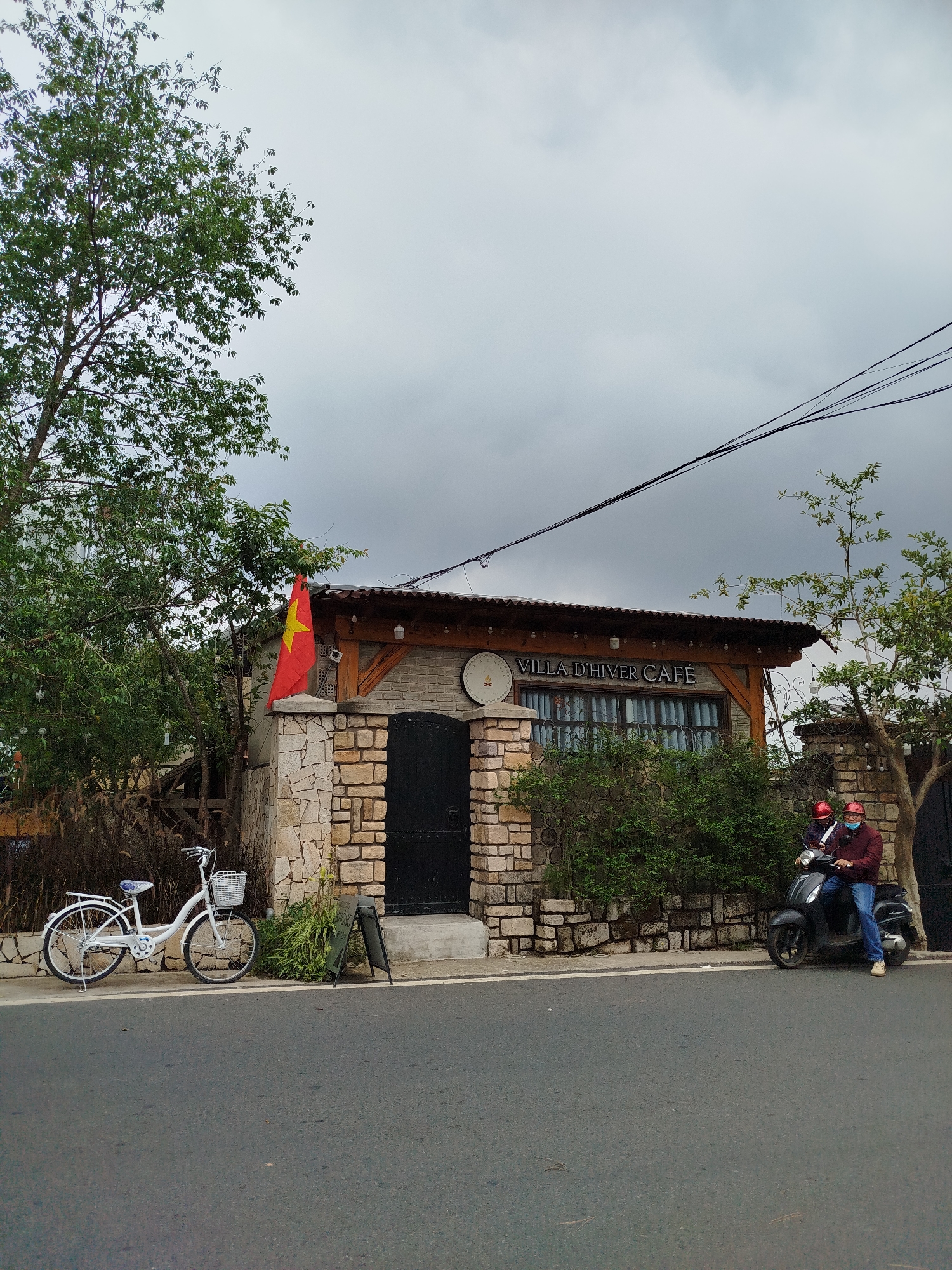 The image captures a roadside scene in Da Lat, Vietnam, featuring a quaint cafe named "VILLA D'HIVER CAFE." The cafe building exhibits a rustic architectural style with stone-clad walls, a dark, arched entrance door, and a wooden beam structure supporting a corrugated metal roof. A red Vietnamese flag with a yellow star is prominently displayed on the left side of the cafe's facade. Decorative string lights are visible under the roofline.
To the left of the cafe, a large tree with abundant green foliage frames the building. The sky above is entirely overcast, indicating a cloudy day, and several power lines crisscross the upper right portion of the frame.
In the foreground, a paved road stretches horizontally. A white vintage-style bicycle with a front basket is parked on the sidewalk near the cafe's entrance, next to a black A-frame sign. Further along the road to the right, two individuals are seated on a dark scooter. The driver wears a red helmet, a face mask, a dark jacket, and blue jeans, while the passenger also wears a red helmet. They appear stationary, possibly having just arrived or paused. The scene depicts a mundane daytime activity under an overcast sky, characteristic of a local establishment integrated into its urban-natural environment.