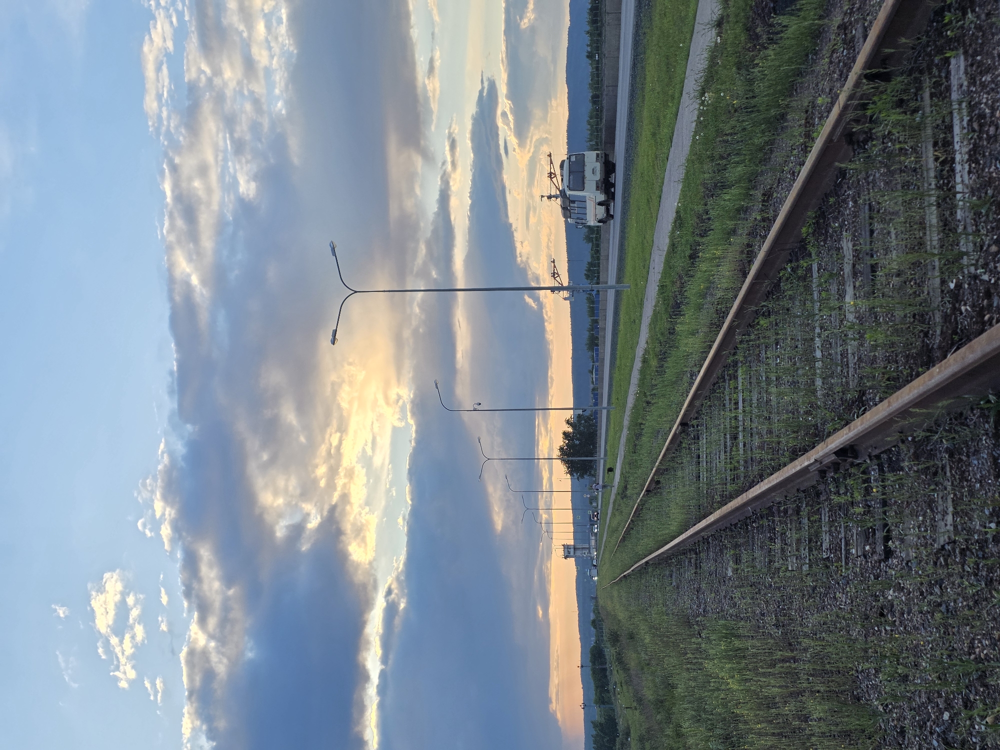 The image shows a view along a railway track in Chaikovsky, Russia. The tracks are overgrown with vegetation. Parallel to the tracks is a road with streetlights and a light-colored utility vehicle parked along it. The sky is visible above, with a mix of blue sky and sunset-colored clouds.