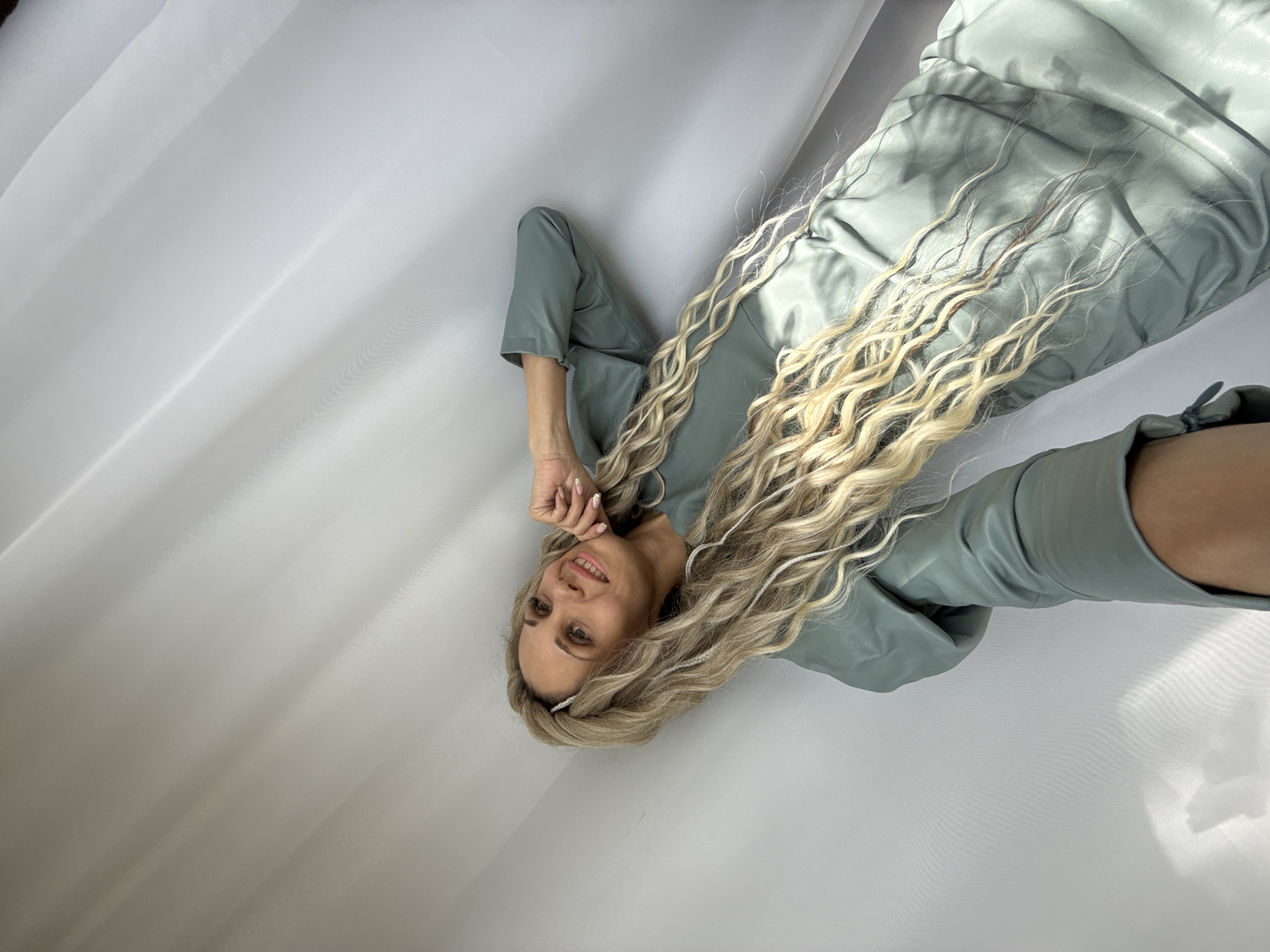 A fair-skinned woman lies on a white surface, possibly a sheet or fabric, in an unknown location in the city of Unknown, Unknown. She wears a light green outfit that includes a top with three-quarter sleeves and knee-length bottoms. Her long, wavy, blonde hair is spread out around her, with some strands appearing to have decorative white ties or ribbons woven into them. She smiles slightly, and her right hand touches her face near her chin. The white surface has folds and appears to be illuminated from above, creating soft shadows.