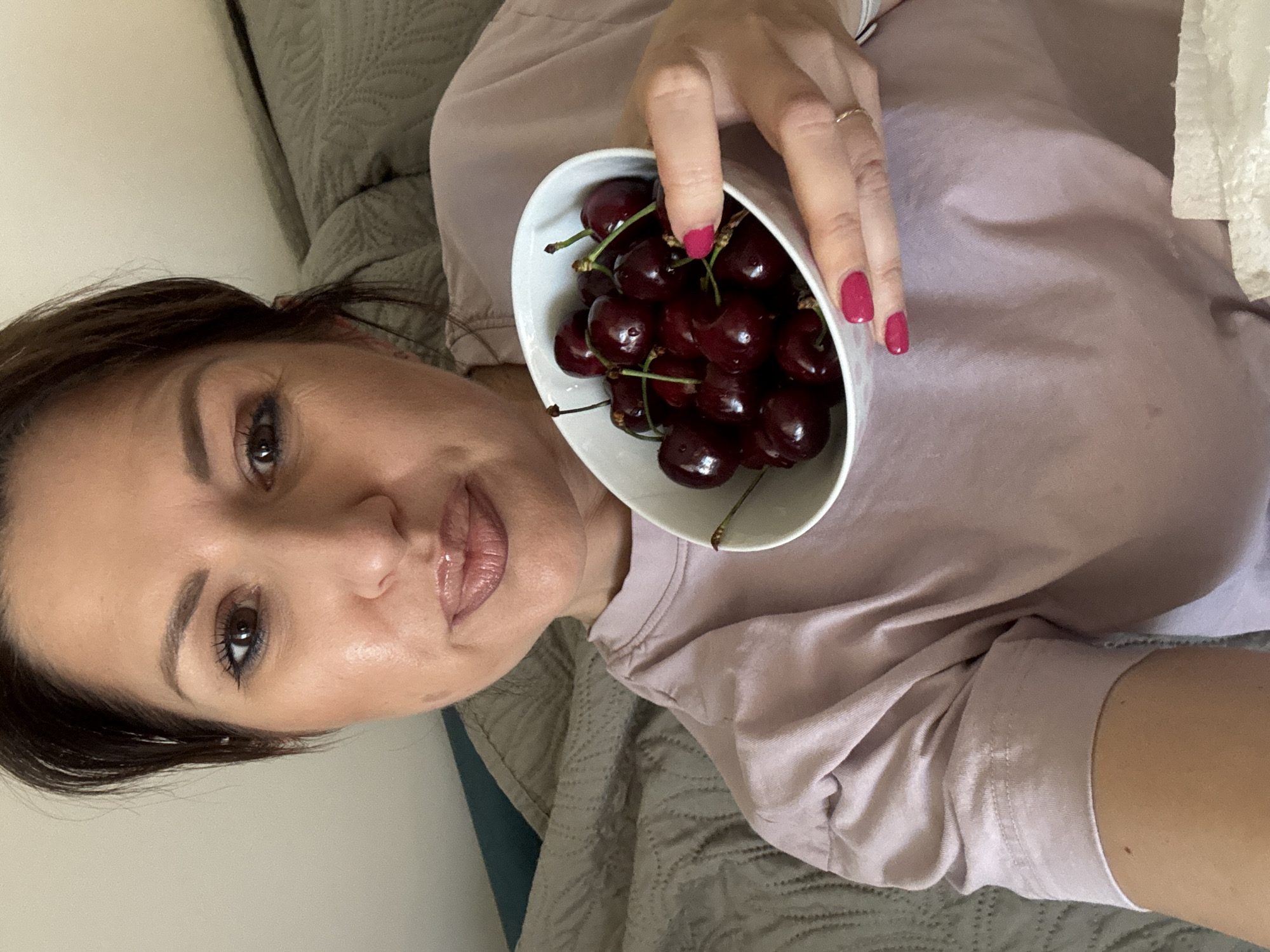 A woman is holding a white bowl filled with dark red cherries, likely in an indoor setting. She is wearing a pinkish-purple t-shirt and has a ring on one of her fingers. Her fingernails are painted a similar shade of pink. The background shows a bed with grey bedding. The photograph is likely taken in portrait mode from a close angle. There is no information given regarding this image being taken in Sofia, Bulgaria.