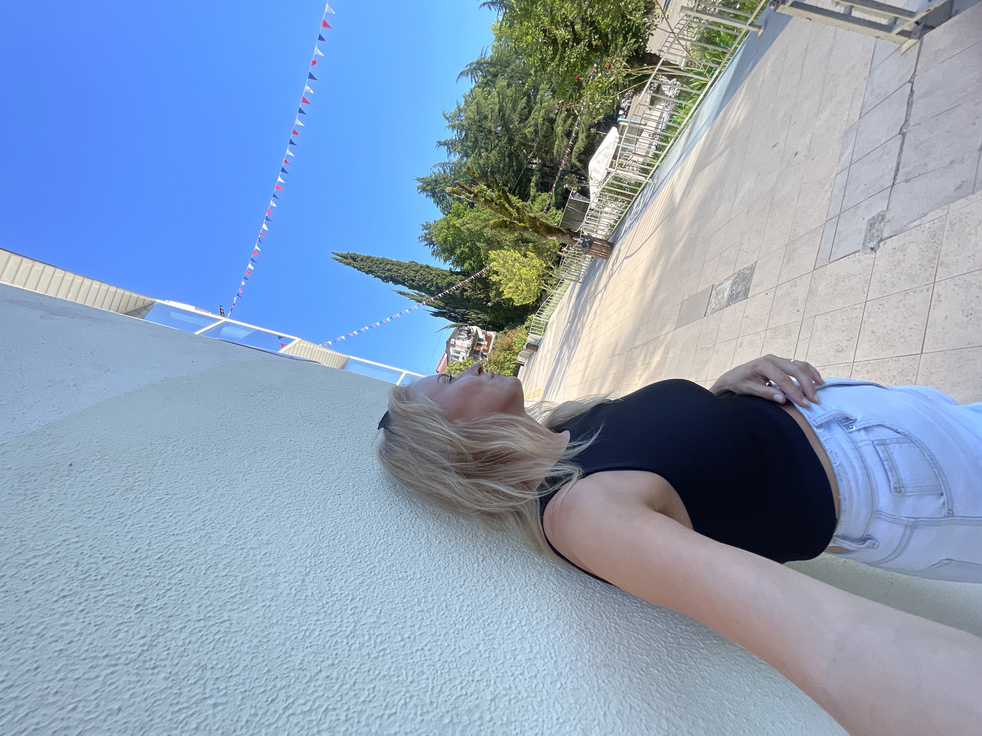 The scene depicts an outdoor promenade or public area, consistent with the subtropical environment of Sochi, Russia, under a clear blue sky. A blonde woman, with sunglasses perched on her head, is captured in profile, leaning against a textured, light beige wall. She is wearing a black ribbed crop top and light-wash denim jeans, with her right hand resting on her waist. Her gaze is directed toward the right side of the frame. The background features a tiled pathway of light-colored squares, extending into the distance, bordered by a white metal railing. Beyond the railing, numerous green trees are visible, including several tall, slender cypress trees, along with distant buildings on a rising terrain. Strings of red, white, and blue triangular bunting are suspended across the upper portion of the frame against the blue sky. A dark-colored cylindrical trash receptacle is situated on the paved area next to the railing. The scene is brightly illuminated by direct sunlight.