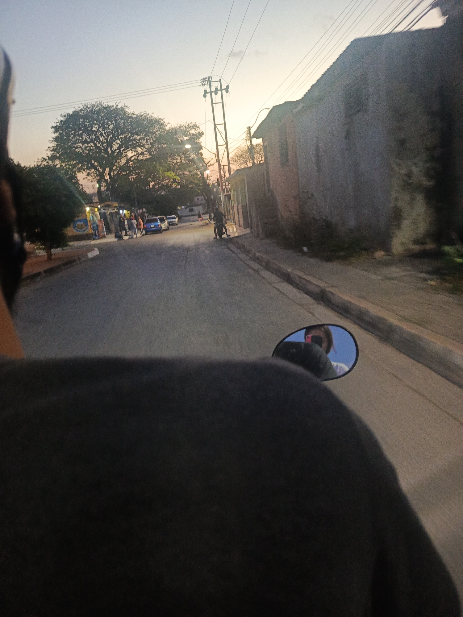 The image captures a first-person perspective from a moving vehicle, likely a motorcycle or scooter, at dusk in Juan Griego, Venezuela. The dark silhouette of a person's back, wearing a black top, occupies the lower foreground, suggesting they are the driver or passenger. A circular rearview mirror on the right side reflects the face and upper body of a light-skinned person, possibly with fair hair, wearing a white shirt and what appears to be sunglasses or a cap, looking towards the camera.
The street ahead, made of asphalt, stretches into the distance, appearing relatively empty save for some activity on the left. On the left side of the street, a group of people is visible near what appears to be a commercial building with a colorful, partially obscured sign that has blue and white elements. A few cars, including a blue sedan and a white car, are parked or moving slowly. Large, leafy trees line this side of the road, and an intricate network of utility poles and overhead wires crisscrosses the sky.
On the right side of the street, an old, weathered building with a sloping roof stands prominently, its facade showing signs of age and perhaps neglect. Further down, other structures are visible. The sky transitions from a pale blue at the zenith to soft orange and pink hues near the horizon, indicative of sunset or early twilight. The overall atmosphere is calm and peaceful, suggesting the close of a day in a local town. No legible text is visible on any signs or objects within the frame.