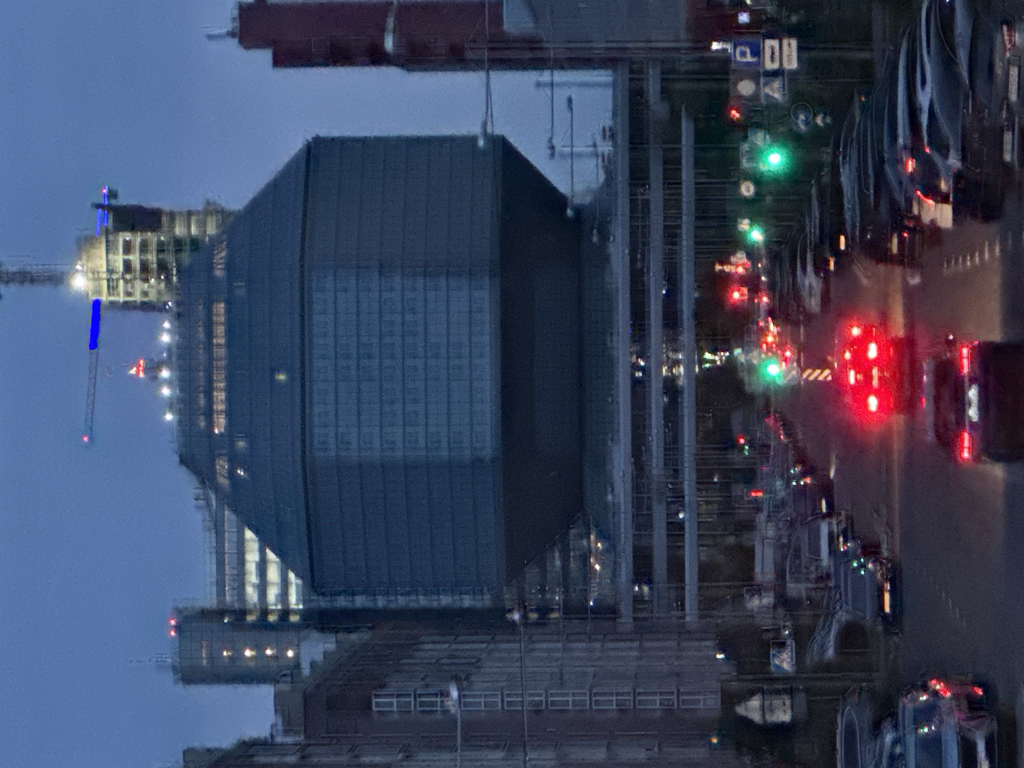 This is a vertical, medium-shot, eye-level, daylight photograph taken in Minsk, Belarus. The photograph captures a view of the National Library of Belarus, a prominent landmark known for its unique diamond-shaped architectural design. The building is the central focus of the image, with its glass facade reflecting the twilight sky. In the foreground, a street is visible with several cars driving on it. The streetlights and traffic lights are illuminated, suggesting it is dusk or dawn. The overall atmosphere of the image is urban and modern, with a blend of contemporary architecture and the hustle and bustle of city life. The photograph is slightly blurred, possibly due to motion or low light conditions, which adds a sense of dynamism to the scene.