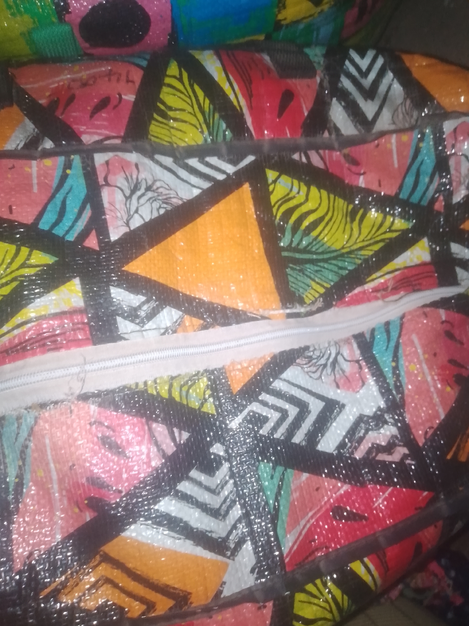 The image displays a close-up of a woven bag with a colorful, abstract geometric pattern. The pattern features triangles and other shapes in shades of pink, yellow, green, turquoise, orange, and black. A visible white zipper runs horizontally across the center of the bag. The texture of the woven material is apparent, and there are areas of shine, suggesting a plastic coating or glossy print. The background is dark and out of focus. There are no people or discernible actions in the image. The context of Jauro Yinu, Nigeria, is not evident from the visual information presented in the photograph.