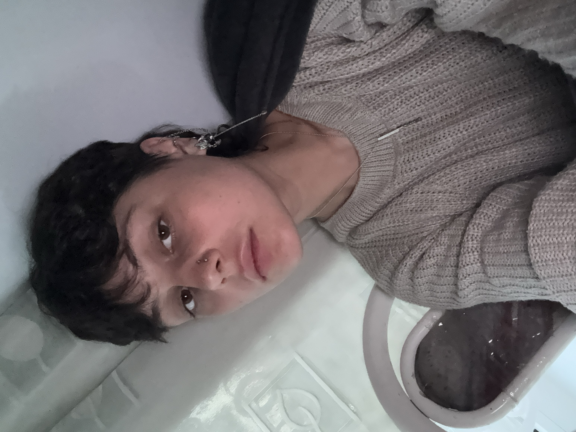 A rotated medium close-up portrait features a young person with dark, wavy hair and fair skin, looking directly at the camera. Their brown eyes meet the viewer's gaze with a neutral, slightly contemplative expression, and their lips are slightly parted. They wear a light brown or beige knitted sweater with a distinct texture, and a dark hooded garment is visible behind their left shoulder and neck.
The person's left ear is adorned with multiple piercings, including a chain connecting two points and several small studs. A thin silver chain necklace with a small, rectangular pendant rests on their sweater. They also have a piercing on their nose, possibly a septum ring or a nostril stud.
The setting appears to be an indoor space, characterized by bright, reflective white surfaces that suggest a bathroom environment. The person seems to be lying or leaning back within a bathtub. In the bottom right corner of the frame, a bathtub drain with a light purple-pink rim is visible, containing dark liquid. Faint raised patterns, resembling leaves or decorative motifs, are subtly embossed on the white surface near the person's head, which could be part of the tub's interior or surrounding tiles. The lighting is soft and even, typical of indoor or artificial light. There is no visible text.