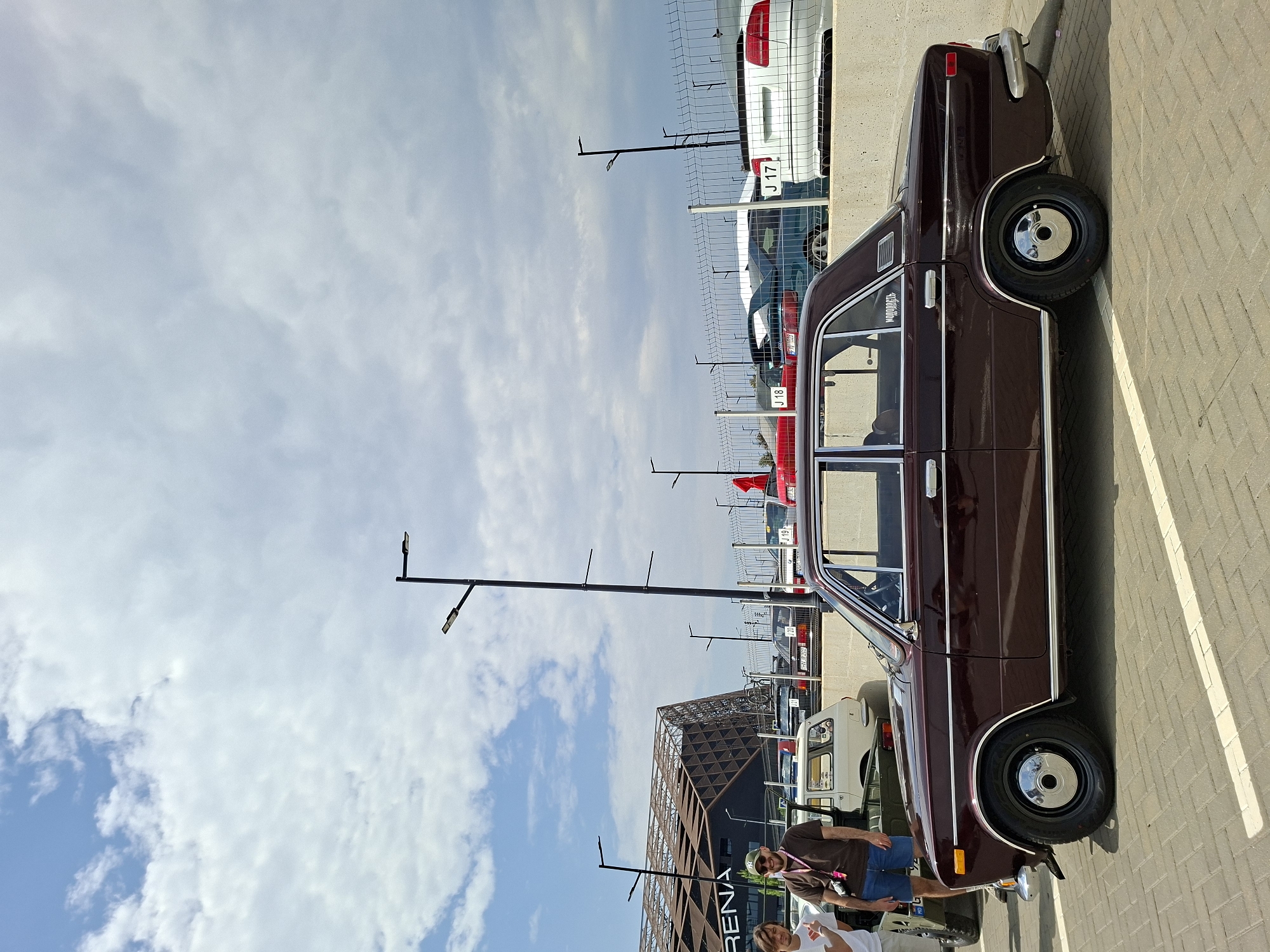 The image captures a scene at the Arena Chișinău, located in Stăuceni, Moldova, during what appears to be a car show or gathering. In the foreground, a classic dark red sedan, likely a Lada, is parked in a paved lot marked with white lines. The car features chrome trim, black wheels with chrome hubcaps, and a sticker on the rear quarter panel reading "Молодость" (Molodost, meaning "Youth").
To the left of the car, two individuals are visible. One man, wearing a brown t-shirt and blue shorts, sports sunglasses and a green baseball cap. He is partially obscured by the car. Another person in a white t-shirt stands next to him.
In the background, behind a fence, several other vehicles are parked, including a white sedan with a license plate that appears to read "J 17", and a red sports car. The Arena Chișinău building, characterized by its modern, angular architecture with a lattice-like facade, is prominent on the left side of the frame. Streetlights are also visible throughout the scene, suggesting an outdoor event. The sky is partly cloudy, with a mix of blue sky and white clouds.