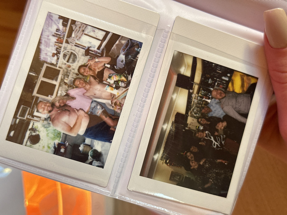 The image displays a close-up of an open white plastic photo album held by a hand with pink manicured nails. The album contains two instant photographs, indicative of Instax or Polaroid film, presented in clear plastic sleeves.
The top photograph, from 3-Y Uchastok, Russia, depicts four individuals posing in what appears to be an outdoor or semi-outdoor kitchen setting. On the far left, a shirtless, muscular man with a beard and short hair, wearing blue shorts or a towel, smiles and gives a thumbs-up with his left hand while embracing a woman to his right. This woman has long blonde hair, wears a light pink short-sleeved top and denim shorts, and has a tattoo visible on her left thigh. To her right, a shirtless man with short hair smiles at the camera. On the far right, a woman with dark hair wears a dark bikini top and makes a peace sign. In the foreground, a table covered with a patterned cloth holds multiple bottles (including beer), a juice carton, and other items. The background reveals a rustic kitchen with white wall cabinets, open shelving filled with various bottles and objects, a round mirror, and a large green watermelon on the floor to the left. The scene suggests a casual gathering.
The bottom photograph, also from 3-Y Uchastok, Russia, shows a group of at least eight adults in what appears to be an indoor bar or restaurant setting. The individuals are clustered together, smiling and laughing. On the far left, a woman in a dark patterned dress smiles. Several women in the center also smile, one wearing a black and white patterned top. To the right, an older man with grey hair in a light blue checkered shirt smiles, standing behind a seated man in a checkered shirt who laughs with an open mouth. The background features a well-stocked bar with numerous bottles on shelves, illuminated from behind. Decorations, possibly streamers, are visible hanging near the ceiling, suggesting a celebratory event.
The immediate surroundings outside the album are out of focus, with an orange-yellow object visible in the blurry background to the left.