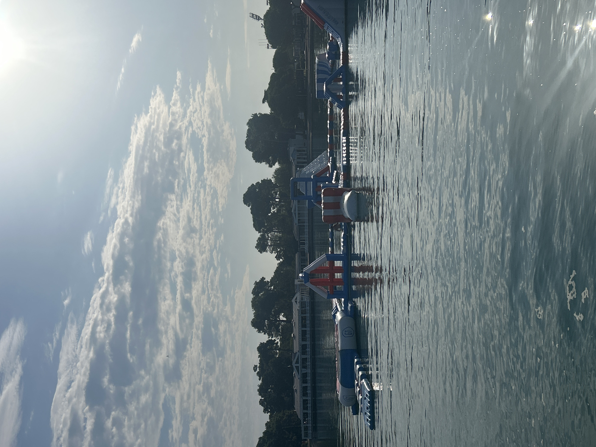 This image shows a bright, sunny day at an inflatable water park located on the water. The park features several inflatable structures, including slides, trampolines, and climbing walls, colored in red, white, and blue. The water park is set against a backdrop of trees and buildings, with a long, white building with a red roof visible on the shore. The sun is shining brightly, creating a glare on the water and casting shadows. The sky is partly cloudy with white cumulus clouds. This scene is located at the beach in Monterol, Croatia.