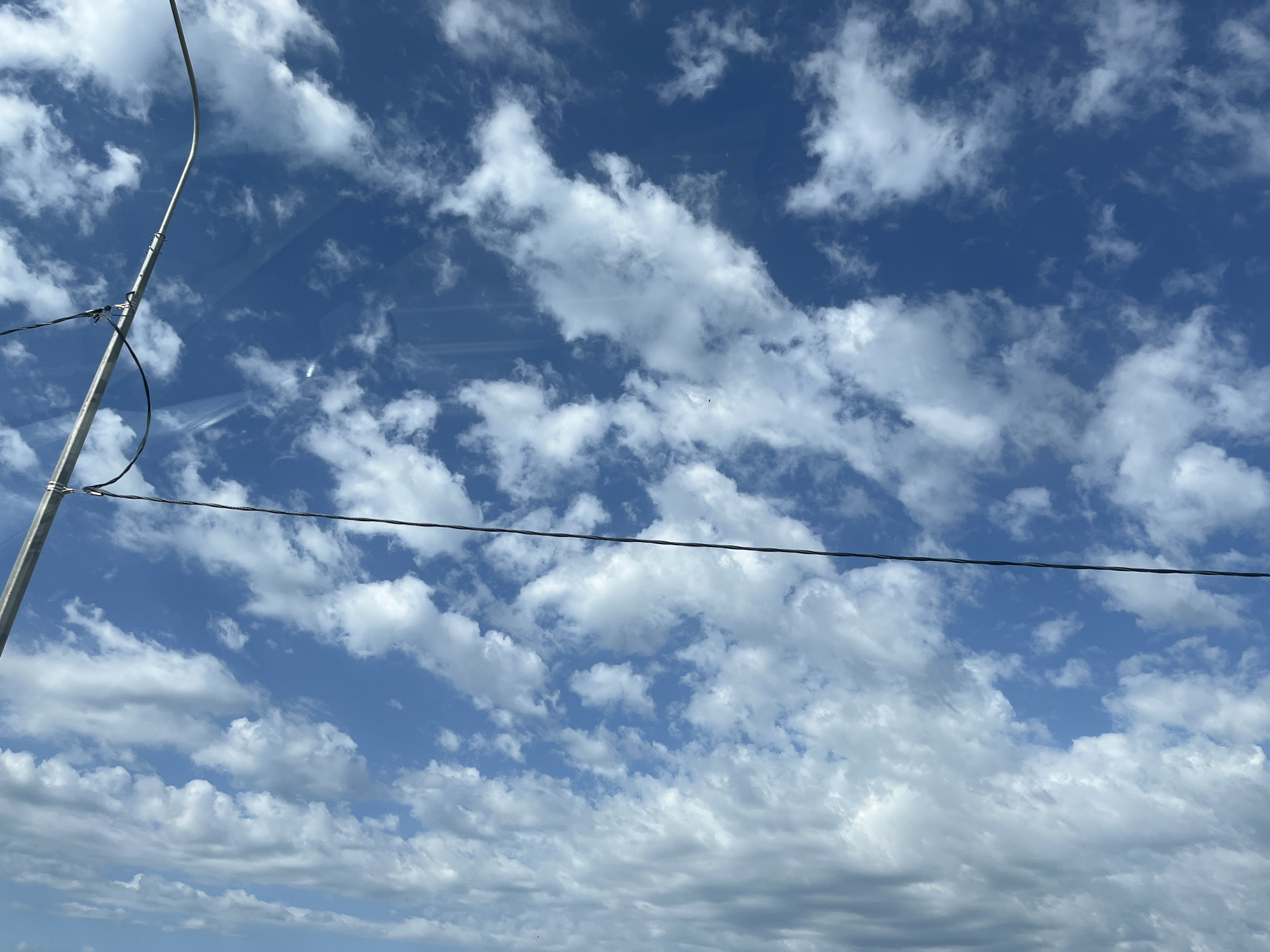 The image shows a sky with a mix of blue and white clouds. A silver pole and wires are visible on the left side of the frame. The location is Tugurgoy, Russia.