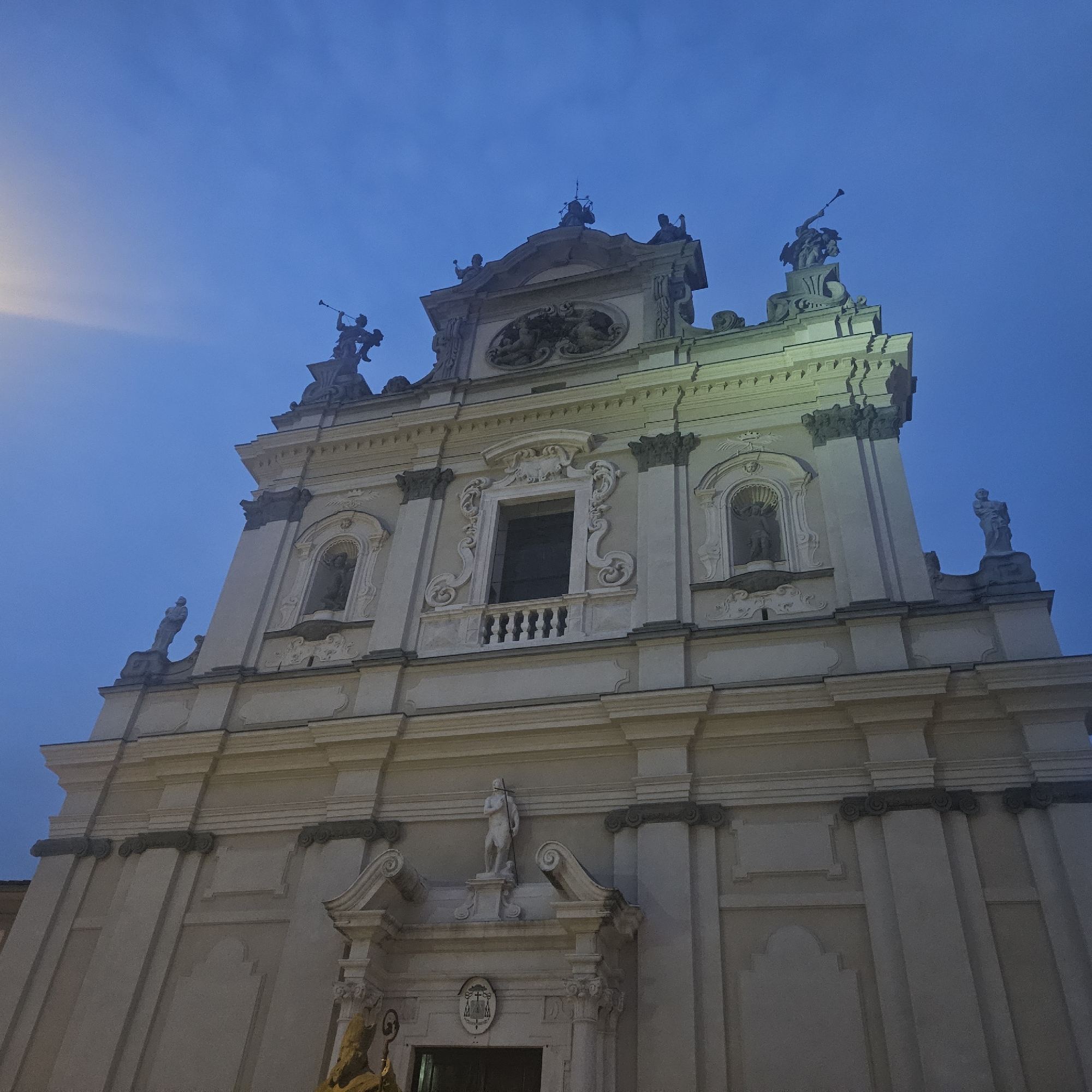 The image depicts the facade of the Parish Church of San Giovanni Battista in Stezzano, Italy, at dusk or night. The church is a multi-tiered structure, with statues adorning the roofline and niches on the upper level. The statues appear to be figures holding trumpets and other symbolic items. The facade is cream-colored, and it is illuminated by artificial lighting, which highlights the architectural details such as the columns and ornate window frames. The sky is a gradient of blue. The main entrance has a statue above it, and architectural embellishments surround the door.