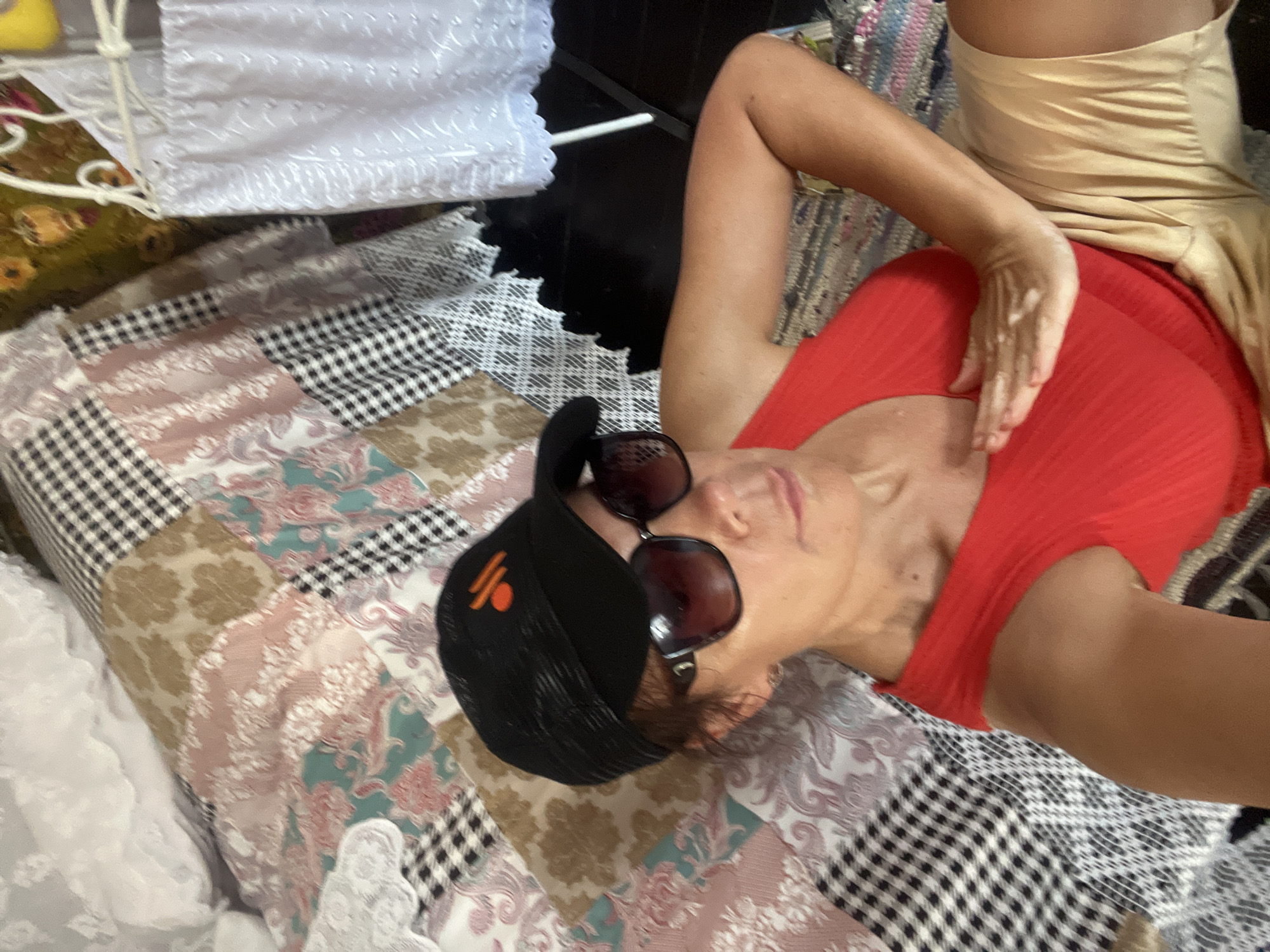 In a dimly lit interior, a woman with brown hair, wearing a black baseball cap, sunglasses, a red tank top, and gold shorts, reclines on a bed covered with a patchwork quilt. Her right hand rests on her chest. The background shows a white lacy canopy over a metal frame. The scene is consistent with an indoor bedroom setting in Taman', Russia.