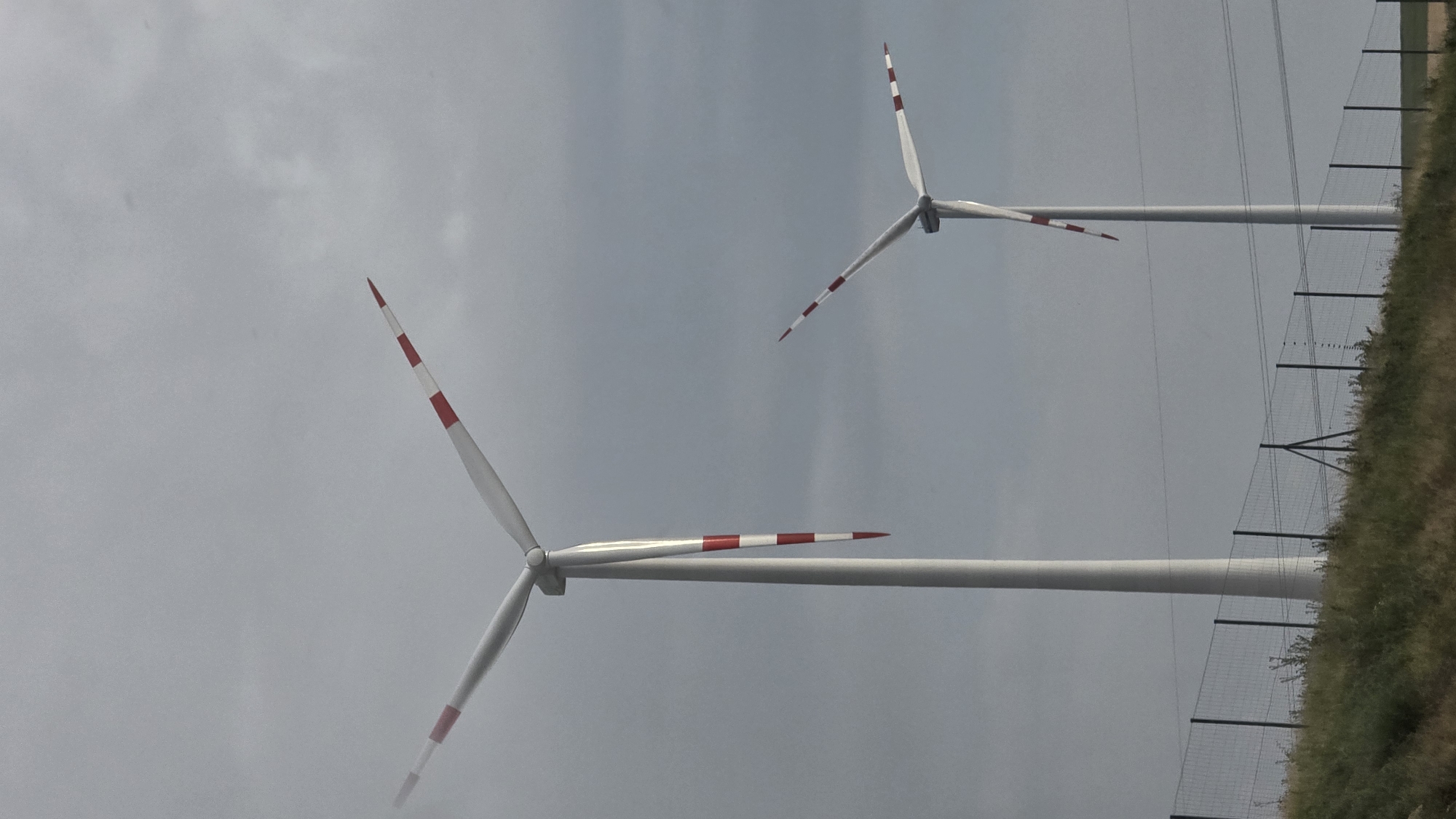 The image shows a view of two wind turbines in Poysdorf, Austria. The sky is overcast and cloudy. The wind turbines are tall, white structures with three blades each. The blades of both turbines have red and white stripes. The foreground consists of a grassy hill with a wire fence along the top. Power lines are visible in the background.