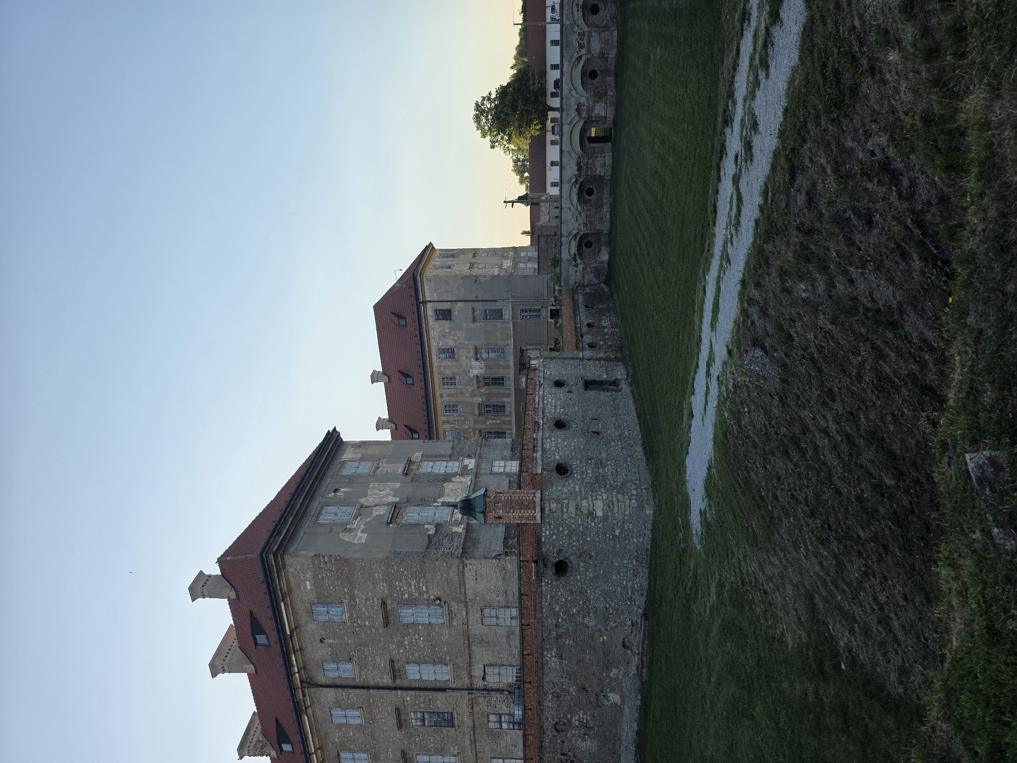 A wide shot of a historic castle structure under a clear, pale blue sky. The castle is a large, aged building with a red-tiled roof and a weathered, grey facade. It has multiple windows, some of which appear to be boarded up or shuttered. A stone wall, topped with a red brick border, surrounds a courtyard area in front of the castle. The courtyard is covered in neatly mown green grass, with a gravel path winding through it. Beyond the castle, a few trees are visible, and in the distance, a white building with a red roof can be seen. The overall lighting suggests it is late afternoon or early evening.