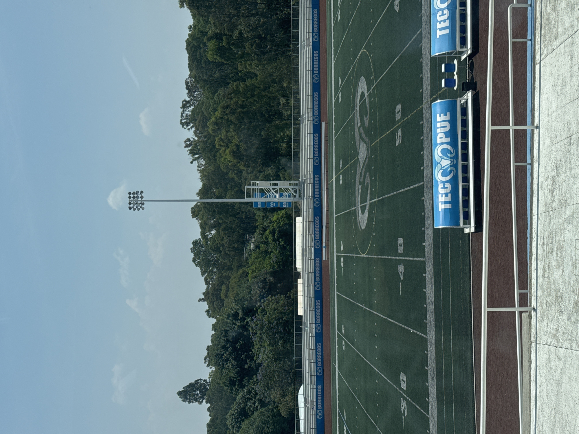 The image presents a sports field, specifically an American football field, located in Puebla, Mexico. The field is marked with white yard lines and features a large logo in the center depicting a ram, the mascot of the Borregos Salvajes, the sports teams of the Tecnológico de Monterrey. The words "TEC PUE" are prominently displayed on blue banners alongside the field, referring to the Tecnológico de Monterrey Puebla campus. Adjacent to the field are stadium seating structures. Beyond the field, a dense forest occupies the background. A tall lighting tower is visible, extending above the treeline. The sky above is pale blue with scattered clouds.