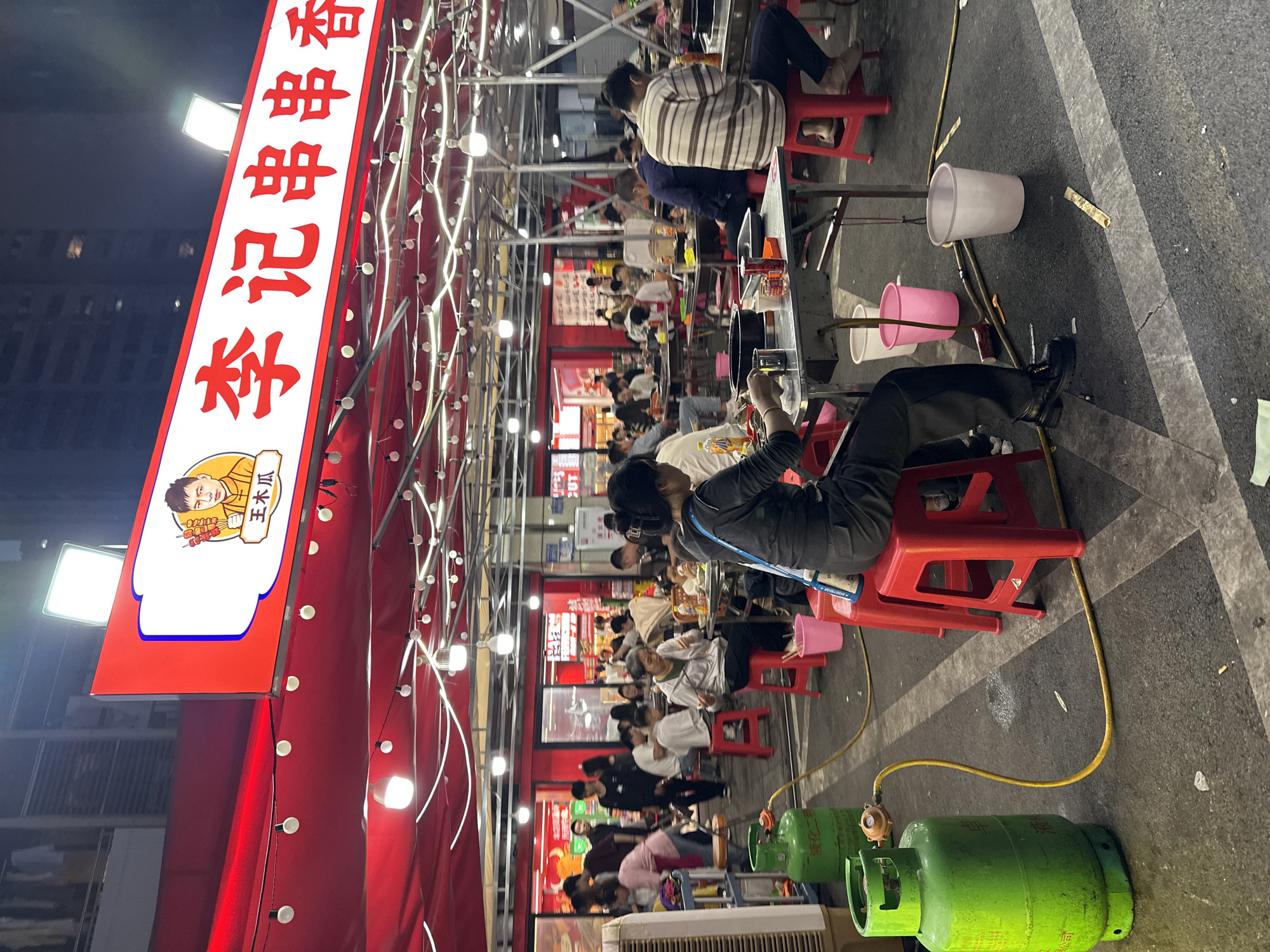 The image depicts an outdoor restaurant in Chongqing High-tech Zone, China, crowded with people dining. Rows of tables and red plastic stools are lined up under a canopy-like structure adorned with lights. Many individuals are seated at the tables, eating and interacting. Gas tanks are visible on the ground, presumably fueling cooking equipment. A large, vertical sign with Chinese characters is prominently displayed.