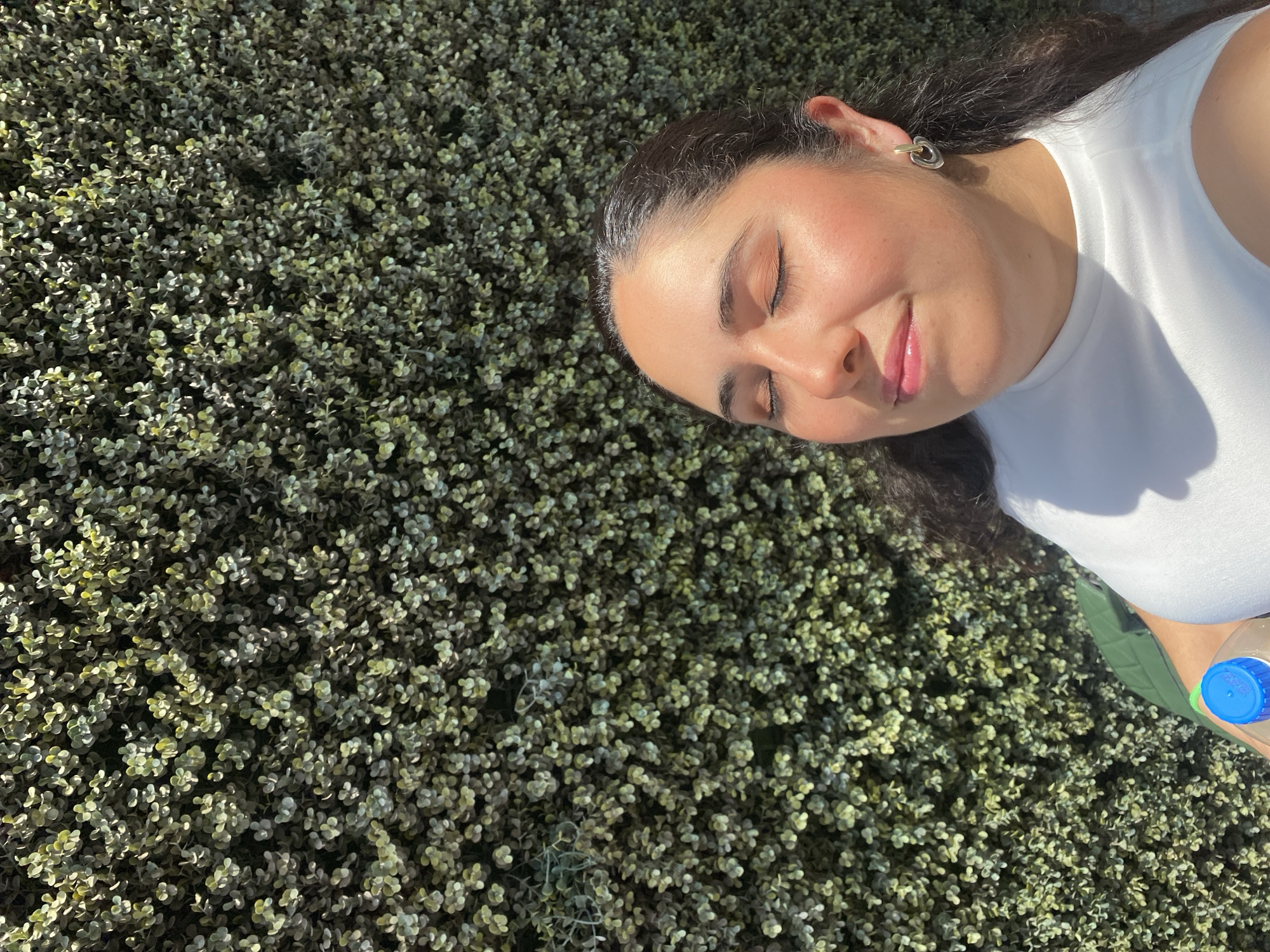 A young woman with dark hair lies on a patch of low-lying, dense green foliage, possibly artificial turf. Her eyes are closed, and she is smiling slightly. She wears a white sleeveless top and a small earring in her left ear. A blue-capped plastic water bottle rests beside her on the foliage. The scene is bathed in sunlight, suggesting a daytime outdoor setting. The exact location within the city of Puebla, Mexico cannot be determined from the provided image.