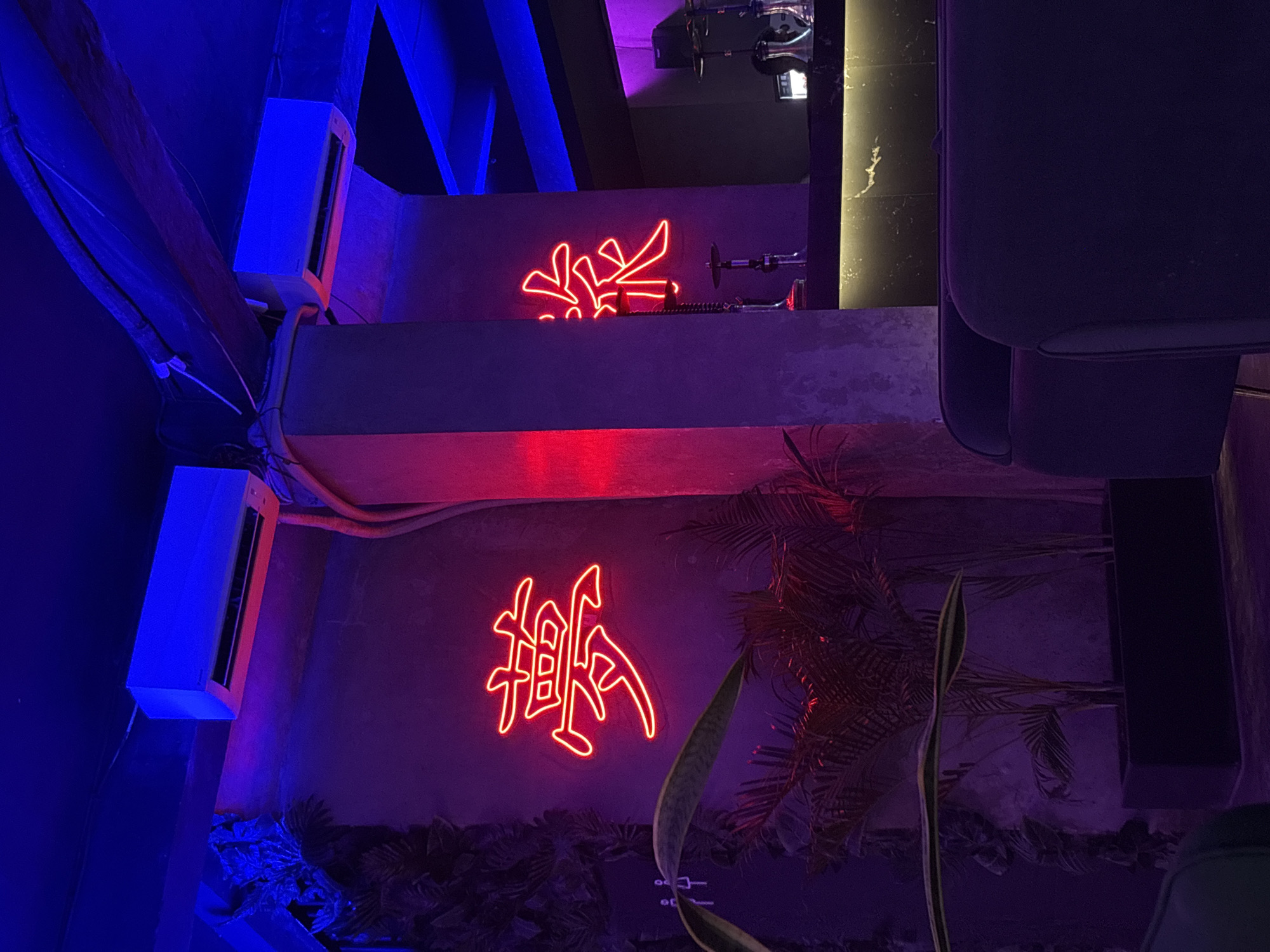 The image depicts an interior setting in Kuta Selatan, Indonesia. Two white air conditioning units are mounted on the left. Red neon signs in the shape of Chinese characters are affixed to the walls. Above one neon sign, a hookah is visible. The lower section of the image shows tropical foliage. Furniture, possibly chairs, are on the right side. The lighting is a mix of blue and red hues.