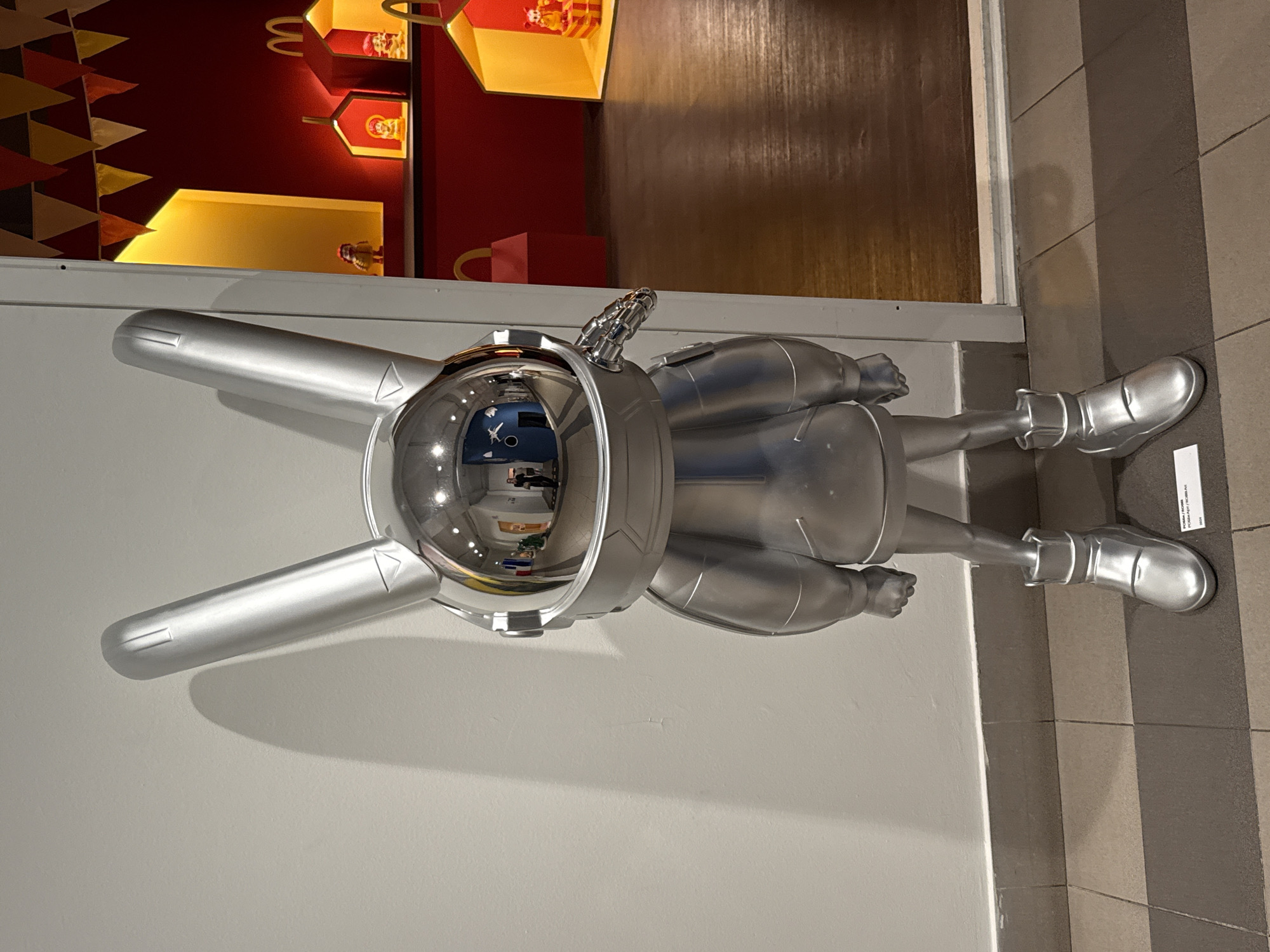 A full-body, straight-on shot shows a large, silver, anthropomorphic rabbit sculpture in an indoor setting. The sculpture is characterized by a reflective, helmet-like head with large, upright ears, a rounded body resembling a jumpsuit, and chunky boots. The helmet reflects the environment, showing a hallway with a person in a dark outfit standing and looking towards the viewer. The sculpture is positioned in front of a plain, light-colored wall. To the right of the sculpture, the scene opens to a display area with a red background and yellow illuminated niches. These niches contain small, colorful figures, some appearing to be toy houses with figures inside, and one displaying a McDonald's logo. A string of triangular, multi-colored flags hangs across the top of this display area. The floor in front of the sculpture is tiled with alternating dark and light grey squares, while the floor in the display area is wooden. A small white label is visible on the floor near the base of the sculpture. The image lacks any clear indicators of its specific location within Moscow, Russia, or any other city.