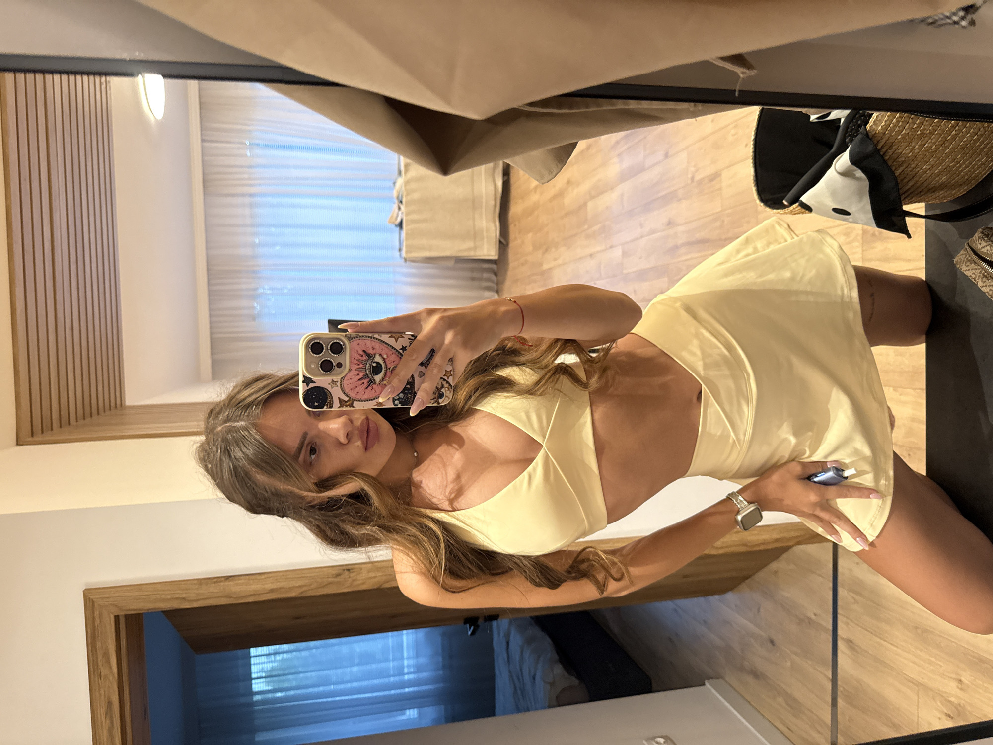 This is a media file of a young woman taking a mirror selfie. She is wearing a light yellow cutout dress and a silver watch on her left wrist. Her phone, with a pink case featuring an eye design, is visible in her hand as she takes the photo. The setting appears to be indoors, possibly a hotel room or apartment, with wooden floors and doors. In the background, there are blinds covering a window, and a straw bag is visible on a surface. The image is oriented vertically, suggesting it was taken with a smartphone. The woman has long, wavy brown hair and is looking directly at the camera. The overall impression is casual and personal, typical of a social media post. The time of day and weather are not discernible. The location is Plovdiv, Bulgaria. There is no visible text in the image.