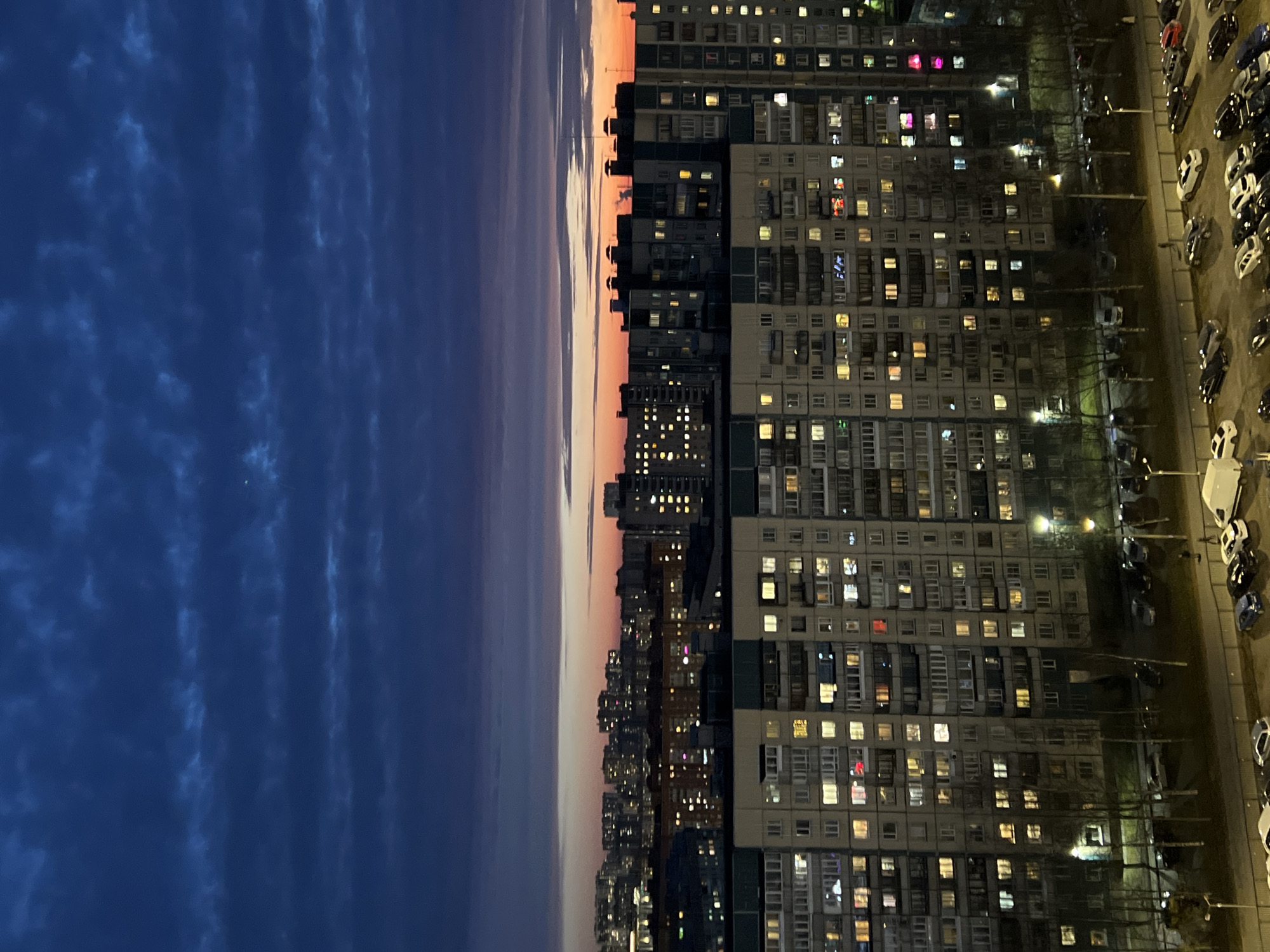The image presents an evening cityscape in Saint Petersburg, Russia. The right portion shows a dense cluster of multi-story apartment buildings, with illuminated windows indicating occupancy. Streetlights cast pools of light on the street-level environment where cars are parked in an orderly fashion. The sky transitions from a twilight hue along the horizon to a dark blue, cloud-streaked sky on the left. This image presents an ordinary urban scene at dusk.