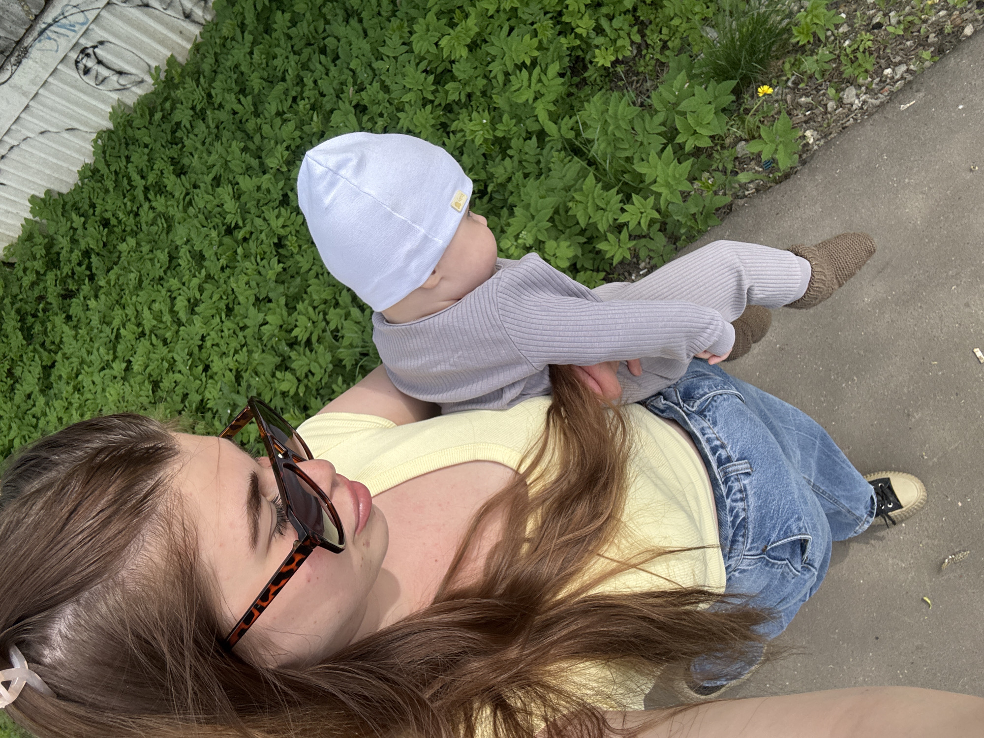 An overhead shot features a woman holding a baby outside. The woman has long brown hair and is wearing sunglasses, a yellow shirt, and blue jeans. The baby is wearing a white hat, a gray onesie, and brown knitted booties. They are standing next to a patch of green foliage and a concrete walkway. The location is in Посёлок комбината стройматериалов-1, Russia.