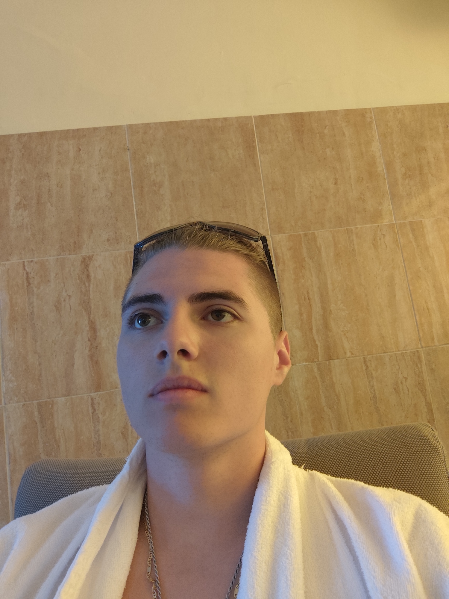 In Hisarya, Bulgaria, a young adult male, appearing to be in his late teens or early twenties, is seated in a chair. He is wearing a white terrycloth robe draped around his shoulders and a silver chain necklace. Sunglasses are perched on top of his head. The background consists of beige tiled walls and a section of lighter colored wall above. The man is looking upwards and to his left, with his facial expression neutral. The lighting suggests an indoor setting, possibly a hotel room or spa.