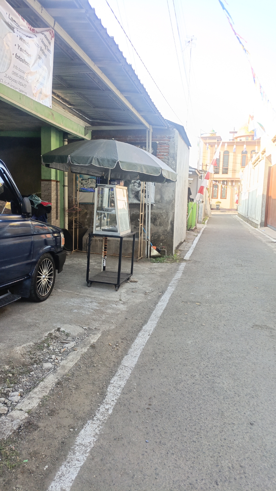 The image depicts a street scene in Salatiga, Indonesia. In the foreground, a dark blue minivan is parked on the left side of the frame. Next to it, there is an outdoor stall with a large dark green umbrella providing shade over a glass display case. The display case is mounted on a black metal stand with wheels. A white line marks the edge of the asphalt road, which extends into the distance. The street is lined with buildings on both sides. On the left, a building with a metal roof and a green-colored lower section is visible, with a white banner hanging from its awning. On the right, a building with yellow and orange accents and a light-colored facade is prominent, adorned with red and white flags. The sky is bright and appears to be overcast or hazy.