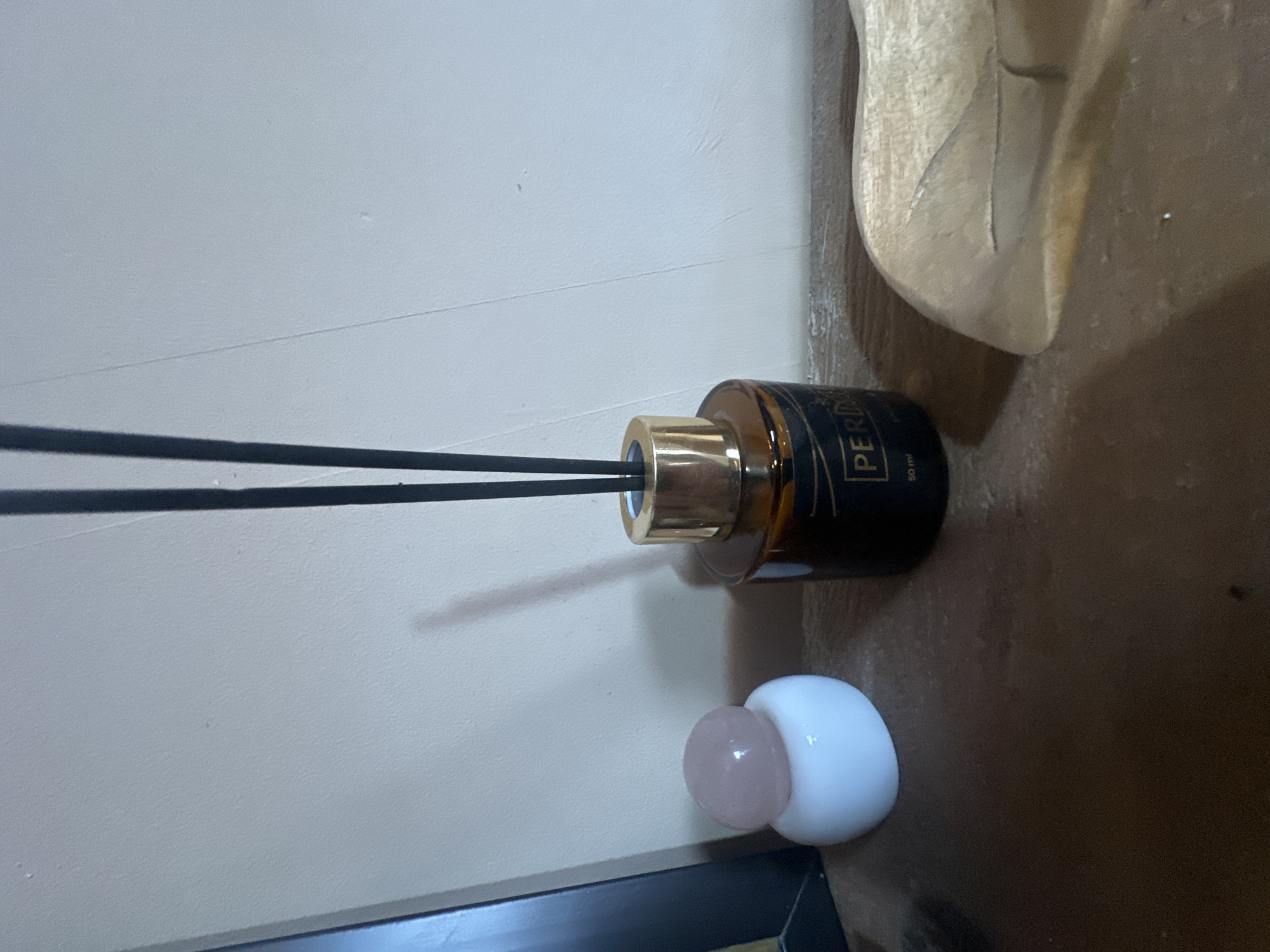 A diffuser with a dark brown glass bottle and a gold colored cap stands on a brown surface. Two black reeds extend upwards from the bottle, one slightly above the other. The bottle has text in gold that reads "PERI... 50 ml". To the right of the diffuser, a pale wooden object with carved details is visible, leaning against the wall. In the foreground, a small, round, white object with a pinkish, pearl-like sphere on top sits next to the diffuser. The background is a light beige wall with some subtle texture and a horizontal line. The scene is indoors, with soft lighting. The overall impression is one of home decor.