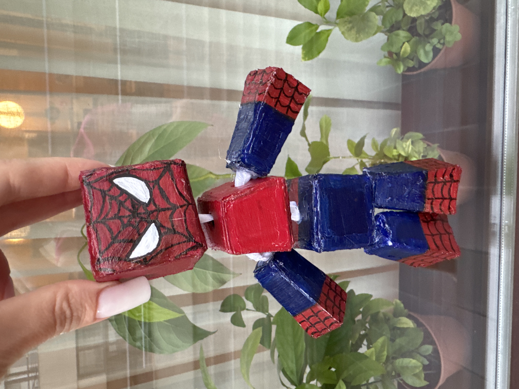 A close-up shot shows a handmade Spiderman figure, constructed from cube-shaped blocks painted red and blue with black web patterns. The figure is held by a person's hand, with fingernails painted in a light pink hue. The Spiderman figure is positioned in front of a blurred background of green potted plants and a sheer curtain, suggesting an indoor setting near a window. The lighting indicates it is daytime. The image is a still frame, with no discernible action or interaction beyond the presentation of the toy. The location is stated as the city of Yalta, Ukraine.
