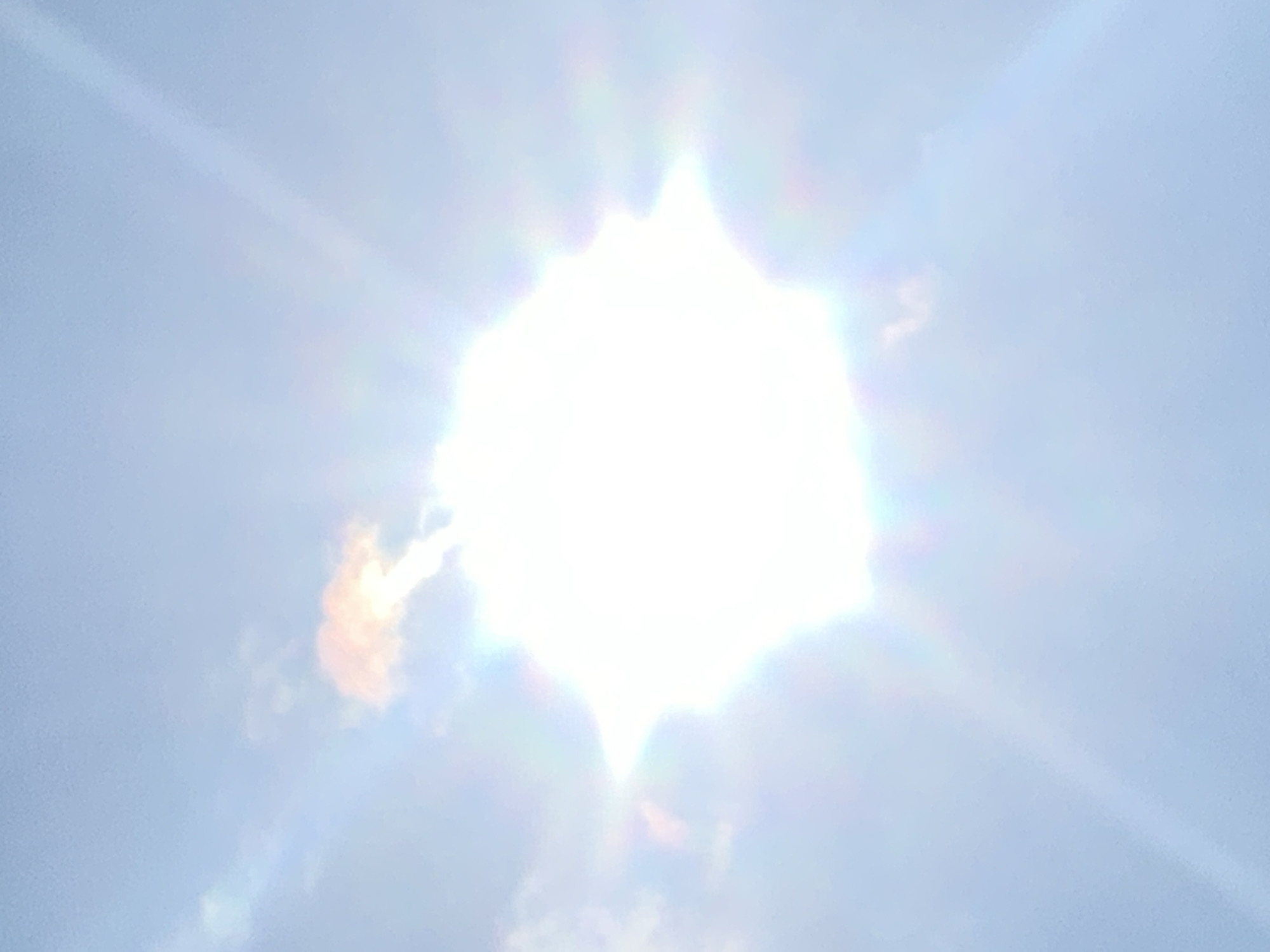 The image shows a bright sun in a light blue sky with some clouds. Sun rays extend outwards from the sun, creating a starburst effect. One cloud appears to have an orange hue, possibly due to the sun's light. There are no visible landmarks that would indicate the specific location within Kolomyia, Ukraine. The scene is a natural, outdoor environment.