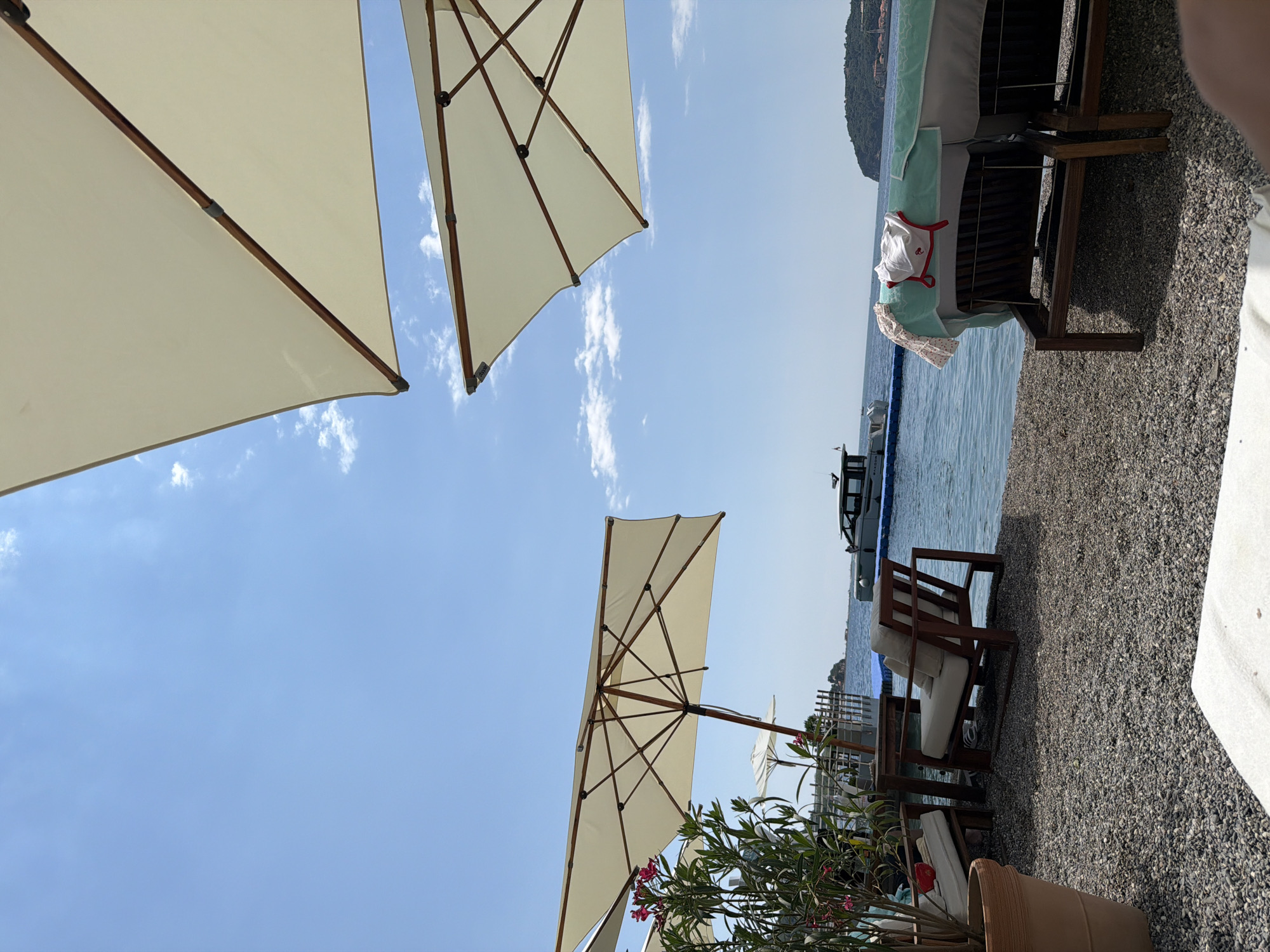 This image depicts an outdoor scene at a beachside resort, likely in Budva, Montenegro. The focus is on a collection of beach umbrellas, providing shade over loungers and chairs. The umbrellas are large and cream-colored, with visible wooden frames. Two of these umbrellas are prominently featured in the foreground, angled upwards towards a clear blue sky dotted with a few wispy white clouds.
In the middle ground, the sparkling blue water of the sea is visible, with a faint outline of a hill or mountains in the distance. Along the shoreline, there are several wooden chairs with white cushions, suggesting a relaxation area. Some items of clothing, possibly a white shirt and a patterned garment, are draped over one of the loungers, hinting at recent activity.
The ground is covered with dark, small pebbles, characteristic of many beaches. A potted plant with pink flowers adds a touch of color and life to the scene. The overall impression is one of a calm and sunny day, ideal for leisure at the seaside. The orientation of the image is such that the sky takes up a significant portion of the frame, emphasizing the bright, open atmosphere. There is no visible text in the image.