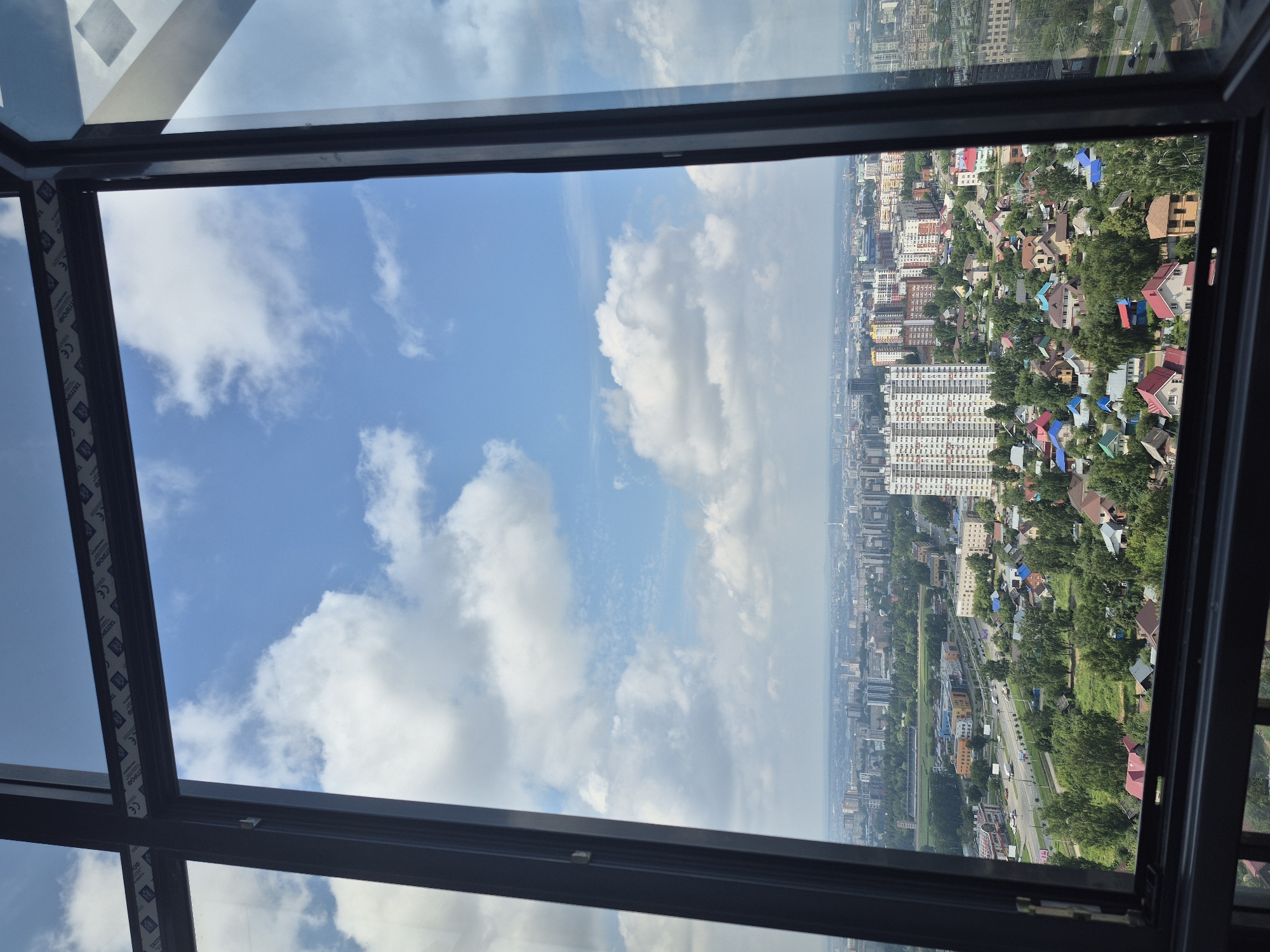 The image is a high-angle view of the city of Izhevsk, Russia, seen through a large, open window. The perspective is from a tall building, looking out over a mixed urban and suburban landscape. In the foreground, the dark frames of the window are prominent, with a portion of the windowpane reflecting the sky.
The mid-ground features a densely populated area with a variety of buildings. A prominent, multi-story white apartment building with many windows stands out in the center of the frame, surrounded by smaller residential buildings with colorful roofs. Trees are scattered throughout this area, interspersed with streets and what appear to be smaller commercial or institutional buildings. A multi-lane road with traffic is visible in the lower left portion of the image.
The background reveals a sprawling city with numerous high-rise buildings, stretching towards the horizon under a bright blue sky filled with fluffy white clouds. The overall impression is of a clear, sunny day in a large Russian city. There are no people or animals visible in the image. The scene is static and appears to be a captured moment of a cityscape.