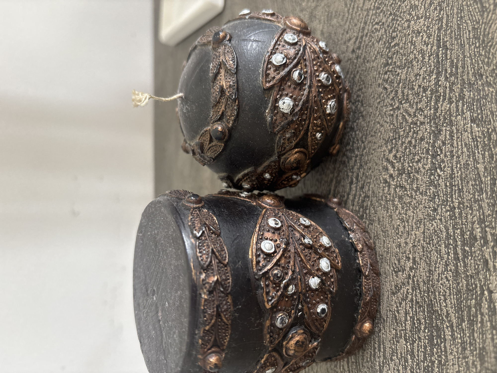 Two dark, ornate candles are displayed on a textured surface. The candle on the left is cylindrical, while the candle on the right is spherical. Both candles are adorned with decorative copper-colored filigree, small clear rhinestones, and what appear to be small dark beads. The spherical candle has a single wick protruding from its top. The background is a plain, light-colored wall. The scene is captured in a close-up shot, focusing on the details of the candles. The image does not provide any information that would allow for identification of a specific city or location, including Dubai, United Arab Emirates.