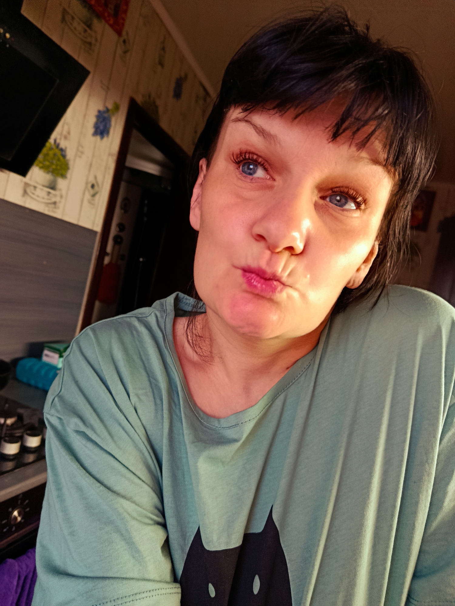 A close-up portrait captures a woman indoors, likely in a kitchen in Фоки, Russia. The woman has short, dark hair with bangs that frame her face and bright blue eyes. Her skin appears fair, and she is illuminated by warm, direct sunlight coming from the right, highlighting her features. She wears a light teal or sage green t-shirt with a black silhouette of a cat visible on the front. Her expression is playful, with her lips pursed as if making a kissy face, and her gaze directed slightly upwards and to her left.
The background reveals a domestic kitchen setting. To the left, a black range hood is partially visible above a counter. The wall behind her is adorned with light-colored wallpaper featuring floral patterns of blue and yellow flowers in vases. Further back, a dark doorway or opening can be seen. On the counter, several small containers and a blue cylindrical object are present, while a control knob from a kitchen appliance, possibly an oven, is visible at the bottom left. No visible text is present in the image. The scene conveys a candid and cheerful moment within a home.