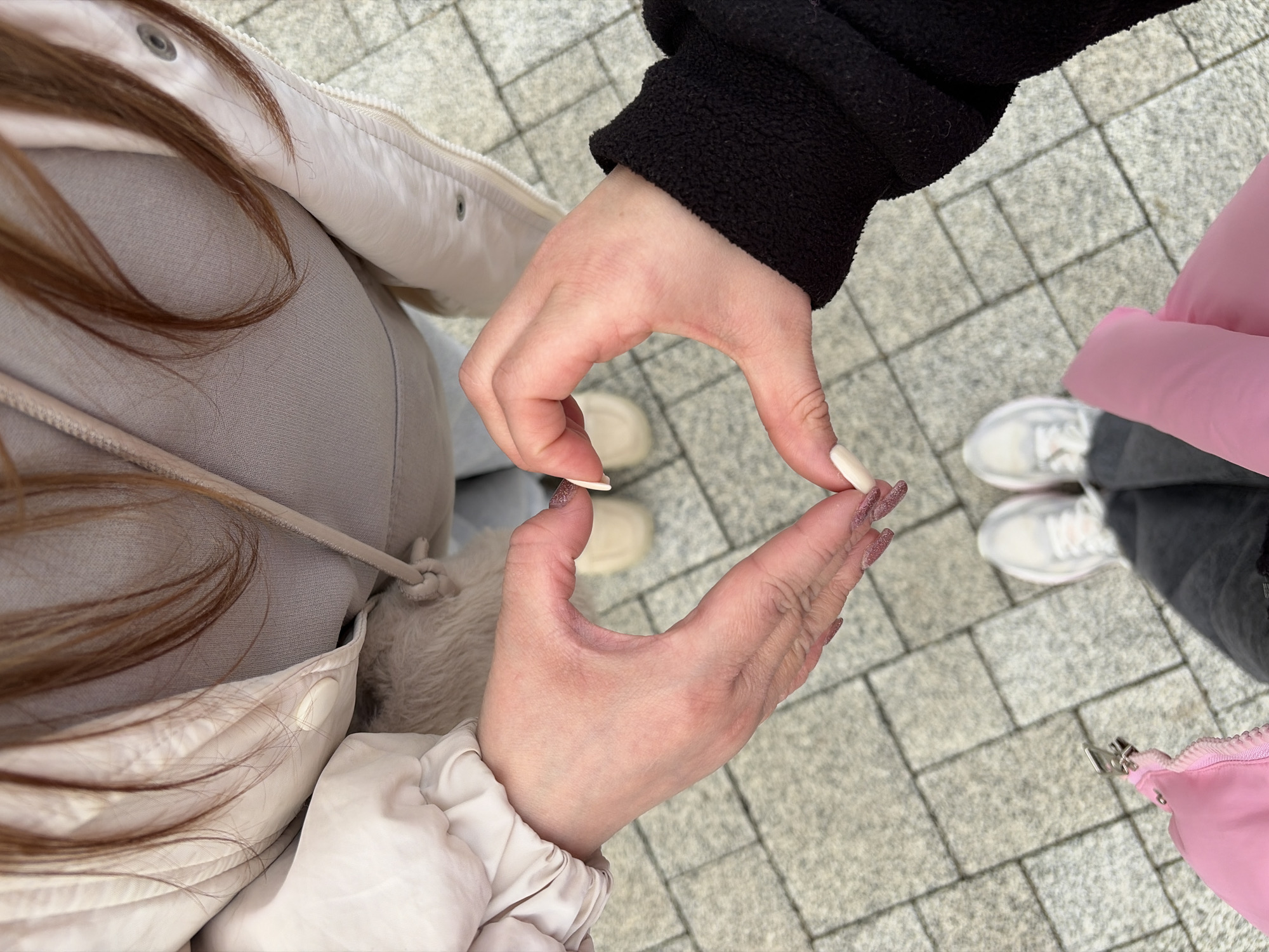 The image presents a top-down view of two individuals standing on a paved surface, likely a sidewalk. The paved area consists of gray rectangular tiles arranged in a repeating pattern.
One person wears a light-colored jacket over a gray top, while the other wears a black jacket. Their hands are positioned to form a heart shape. The person in the light-colored jacket has long, decorated nails.
Their feet are visible: the individual in the light jacket is wearing beige shoes, while the other is wearing white sneakers. A pink fanny pack or small bag is also visible near the person in the black jacket.
There is no indication of the location being Novopokrovskaya, Russia.