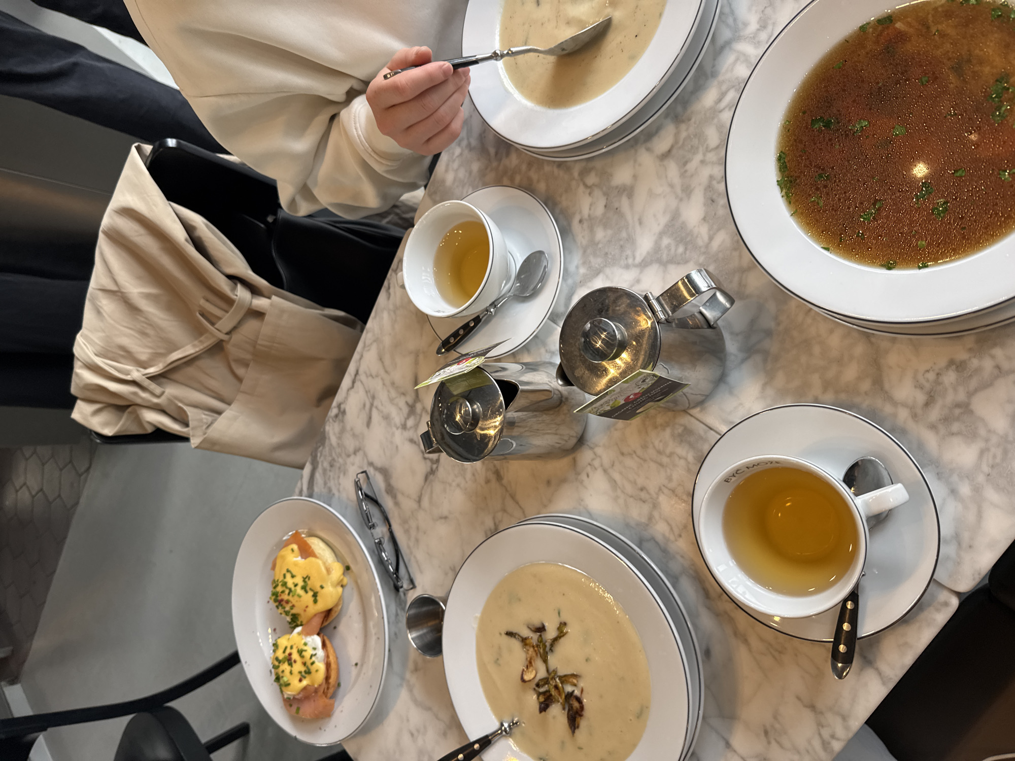 The image depicts an aerial view of a dining table at a restaurant or café. The table, likely in Warsaw, Poland, features a marble top. On the table are the following:
1. A person is visible, wearing a light-colored long-sleeved garment, using a spoon to eat soup.
2. Two bowls of soup, one being cream-based with mushroom garnish, and the other a clear broth with herbs.
3. A plate of Eggs Benedict.
4. Two teacups with saucers, containing a light-colored beverage, presumably tea.
5. A tea pot.
6. Flatware including spoons.
A handbag is visible on a chair beside the table. The setting suggests a casual yet refined dining experience.