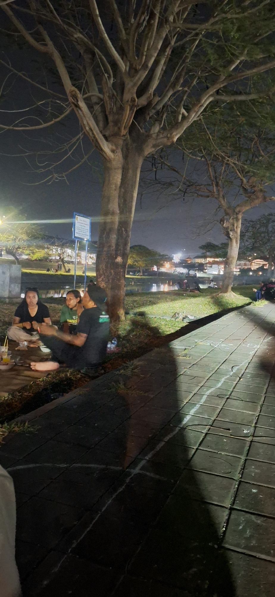 The image depicts an outdoor scene at night, likely in Denpasar, Indonesia. In the foreground, three individuals are seated on the ground near the base of a large tree. One person, wearing a black t-shirt and pants, is facing away from the camera and appears to be interacting with someone to their right. Another person, wearing a black t-shirt, is seated facing the camera, and a third person, wearing a green t-shirt, is seated between them. There are some disposable cups and what appears to be food items on the ground in front of them. A paved walkway with chalk markings runs diagonally across the right side of the image. In the background, there is a body of water, possibly a canal or river, with lights reflecting on its surface. Further in the distance, buildings and trees are visible, illuminated by artificial lights, suggesting an urban or semi-urban setting. The overall atmosphere is casual and relaxed, with people enjoying an evening gathering.