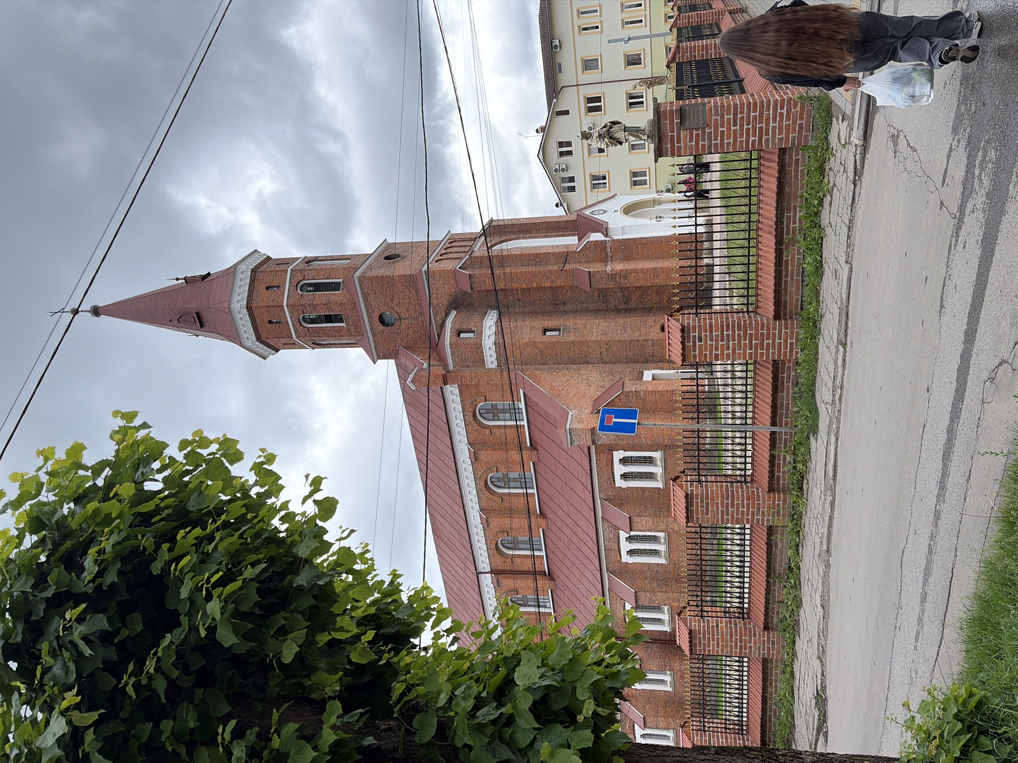 This is a full, eye-level shot of a large, brick church with a red-tiled roof and a tall, red-spired bell tower. The church is located on the left side of the frame, and the street is in the foreground. To the right of the church is a light-colored, multi-story building with many windows. A person with long, brown hair is walking away from the camera on the sidewalk, carrying a white plastic bag in their right hand. There is a black metal fence with gold accents in front of the church. The sky is overcast.