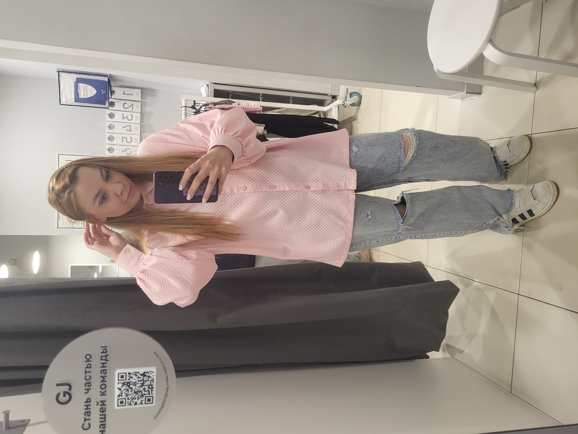 A woman stands in a fitting room at a GJ retail store, located in Chaikovsky, Russia, holding a smartphone to take a selfie in the mirror. She wears a light pink textured, long-sleeved button-down shirt with puffy sleeves and distressed light blue denim jeans, paired with white Adidas sneakers. The fitting room has a gray privacy curtain, a round white stool, and tiled floors. A sign on the curtain invites customers to join the GJ team and includes a QR code. In the background, clothes hang on a rack, and numbered labels are visible on a shelf.