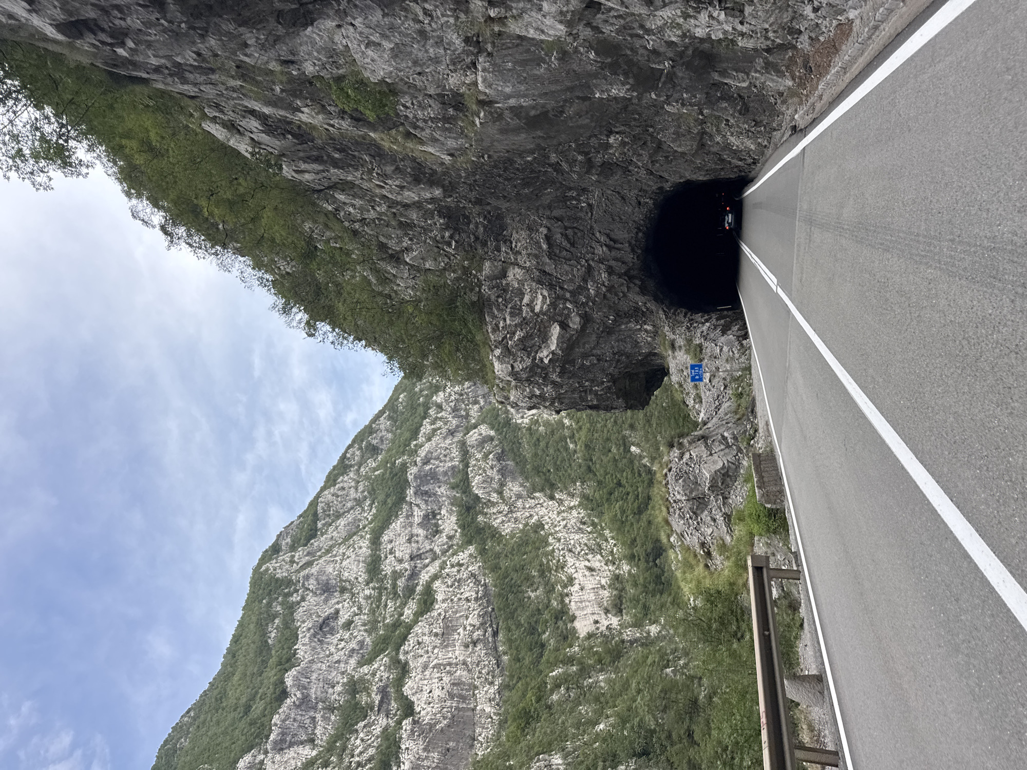 The image shows a two-lane asphalt road in Vlahovići, Montenegro, curving towards a dark tunnel carved into a sheer rock face. The road is bordered by a metal guardrail on the left and a steep, rocky embankment on the right. The mountains surrounding the road are covered with dense green vegetation, with rocky outcrops visible. The sky is partly cloudy. A single white car is visible entering the tunnel. A blue road sign with white text is posted on the left side of the road, near the tunnel entrance. The scene depicts a mountainous landscape with a road leading through a natural rock tunnel.