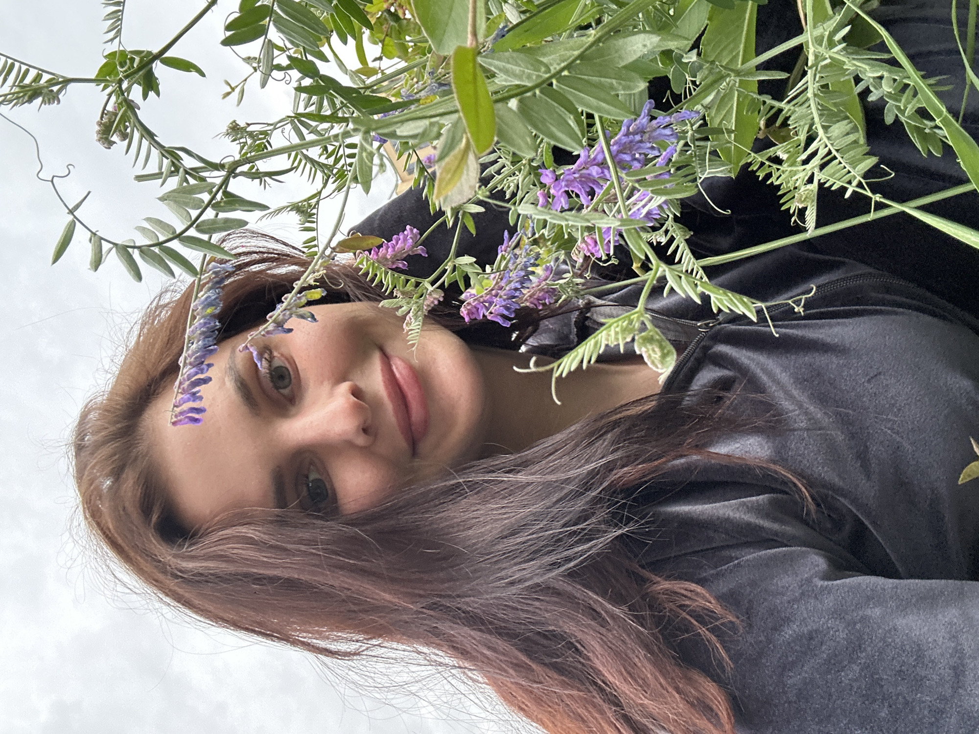 A close-up selfie shows a young woman with long, dark auburn hair and fair skin. She is wearing a dark gray hooded jacket. Her head is tilted slightly to the left, and she is smiling gently at the camera. She has a sprig of small purple flowers resting on her forehead and another sprig near her left eye. The background consists of an overcast sky and lush green foliage with more purple flowers. The image appears to have been taken outdoors in a natural setting.