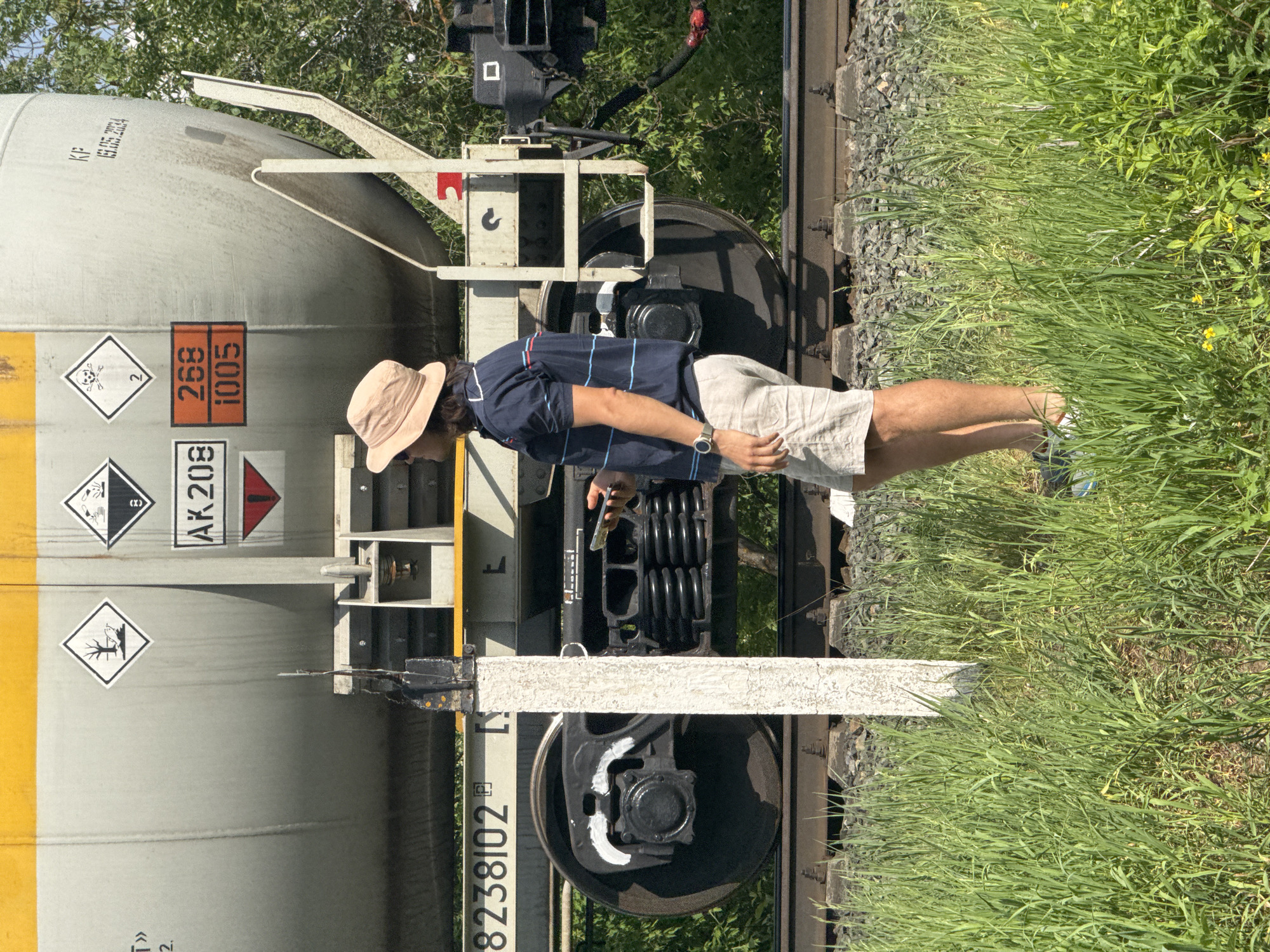 A man wearing a tan hat, navy blue striped polo shirt, and tan shorts is standing beside a rail car on railroad tracks. The rail car is light gray and has orange hazard placards. A signpost is planted between the rails. Tall grass surrounds the tracks. The exact location is unknown; the image does not confirm it is in Izhevsk, Russia.