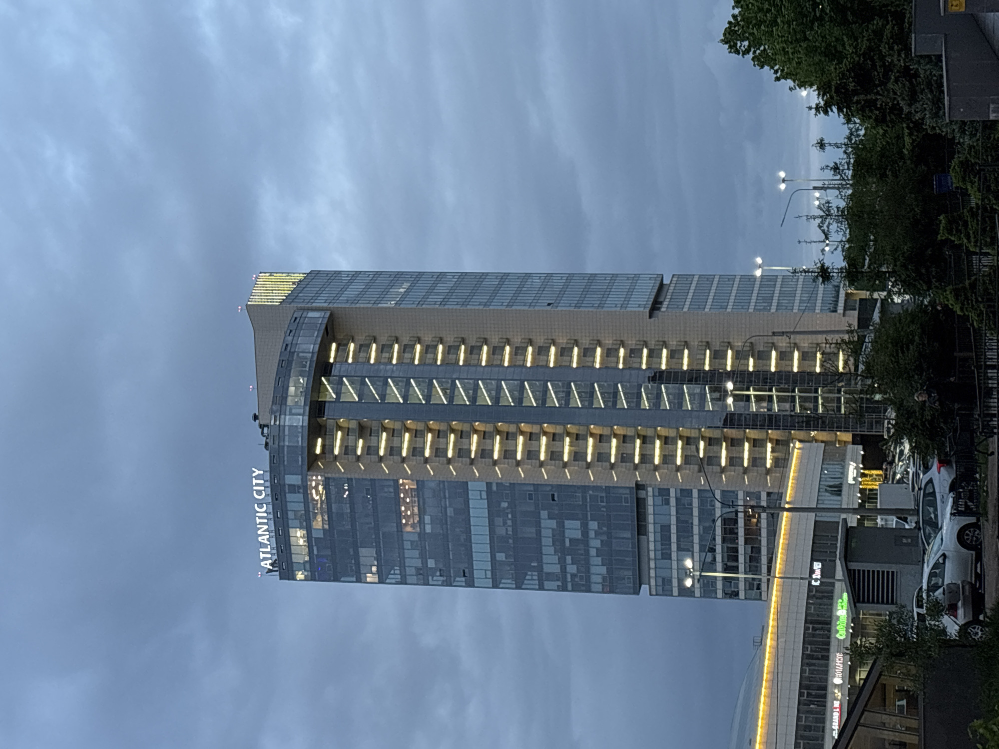 The image shows the Atlantic City business center in Saint Petersburg, Russia. The building is a tall, modern skyscraper with a glass facade. The name "ATLANTIC CITY" is vertically displayed on the side of the building. It is illuminated with rows of lights. The sky is overcast, and the light indicates it may be evening or dusk. There are trees and parked cars in front of the building. Streetlights are also visible. Other commercial buildings and retail spaces are present at the base of the skyscraper.