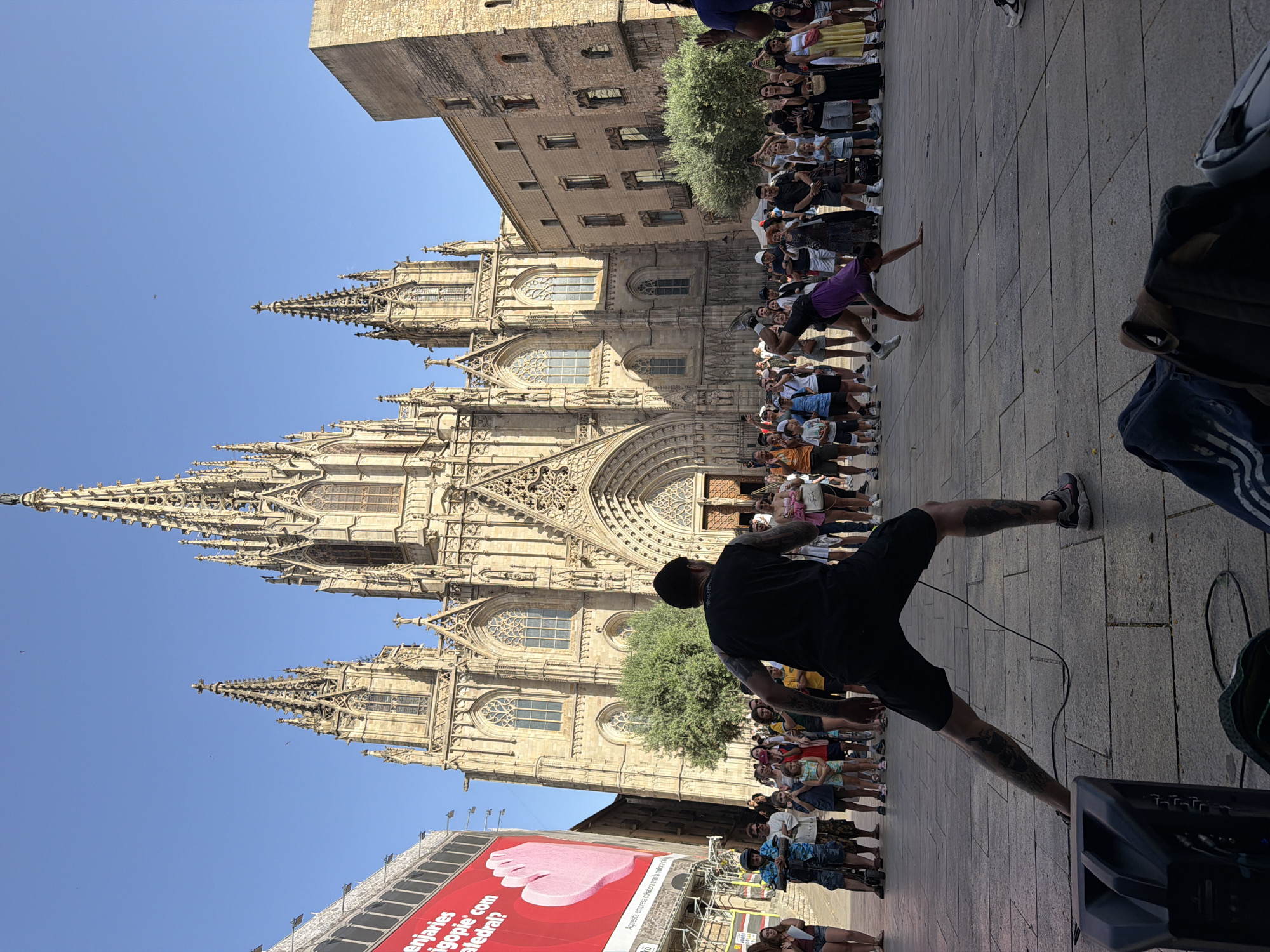 The image depicts a lively street scene in front of the Barcelona Cathedral in Barcelona, Spain. A crowd of people has gathered to watch a performance. In the foreground, a man with extensive tattoos on his legs is facing away from the camera, appearing to be the DJ or MC for the event, as a speaker is placed near his feet. Further into the scene, two performers are engaged in an acrobatic dance. One performer, wearing a purple outfit, is in mid-air, executing a complex move. The other performer, dressed in dark clothing, is also in motion, supporting the airborne performer. The background is dominated by the impressive Gothic architecture of the Barcelona Cathedral, with its intricate spires and facade. Adjacent to the cathedral, a modern building with large windows stands to the right, and a large red billboard advertising an event is visible on the left. The ground is paved with large square stones, and the sky is clear and blue.