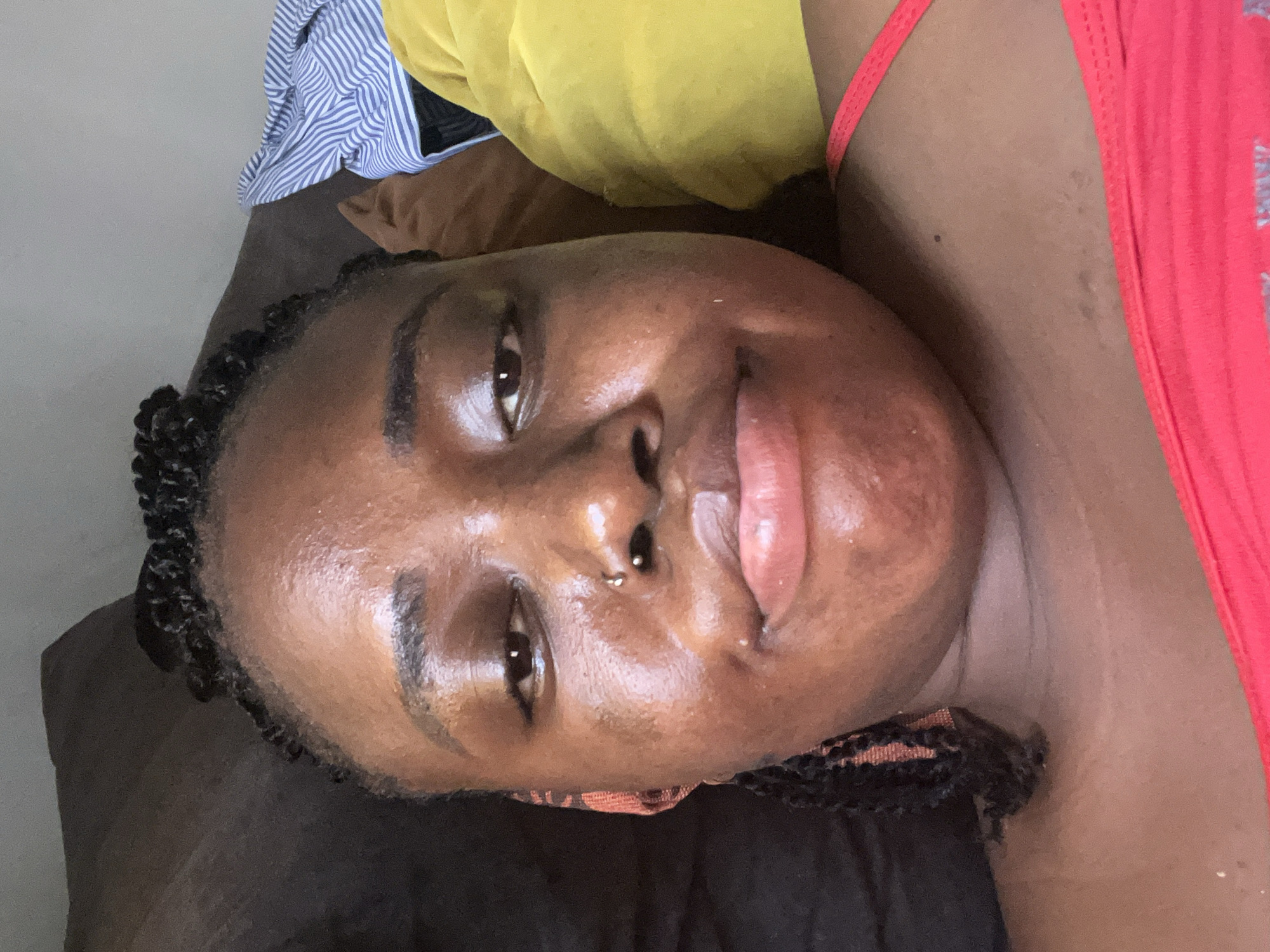 A close-up, over-the-shoulder selfie shows a Black woman lying down, her face filling most of the frame. Her dark hair is styled in braids, with some braids visible framing her face and at the back. She is wearing a bright red halter top. Her skin appears dewy, with a slight sheen. She has a nose ring and is looking towards the camera with a neutral expression. To her right, a yellow pillow and a blue and white striped shirt are visible. The background consists of a dark, textured surface, likely upholstery, which could be a couch or bed. The overall lighting is soft, suggesting an indoor setting. While the image is taken in Abuja, Nigeria, no specific landmarks or identifying environmental features of the city are discernible in the frame.