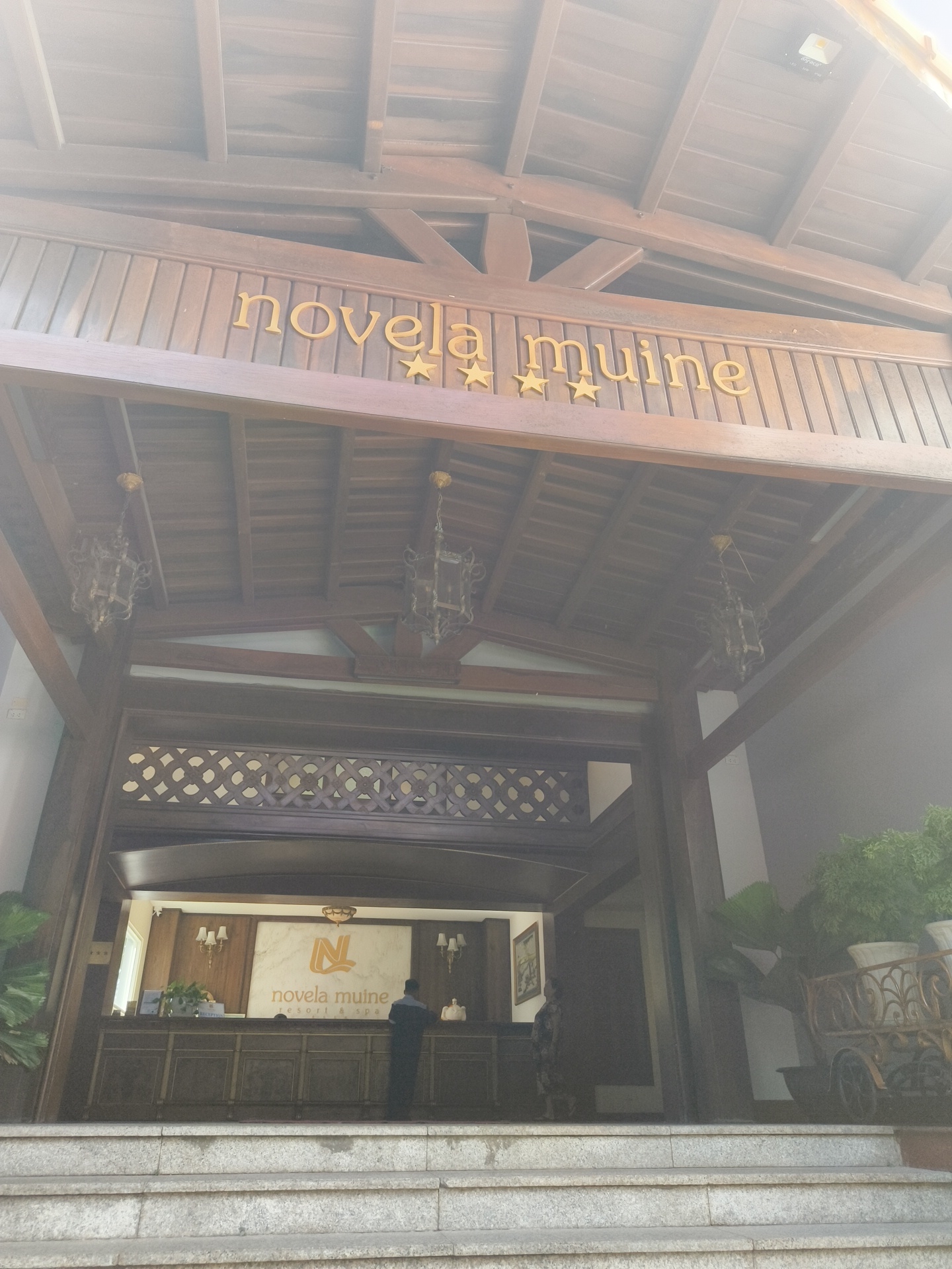 The image shows the entrance to the Novela Muine Resort & Spa in Phu Thuy Ward, Vietnam. The entrance features a wooden awning with the resort's name "novela muine" and three stars spelled out in gold lettering. Two ornate lanterns hang from the ceiling of the awning. Below the awning is the reception area, with a counter and a large sign displaying the resort's logo and name. A person in a dark uniform stands behind the reception counter. The building has a traditional architectural style with wooden beams and decorative latticework. The area in front of the resort has stone steps leading up to the entrance, and potted plants are visible on either side.