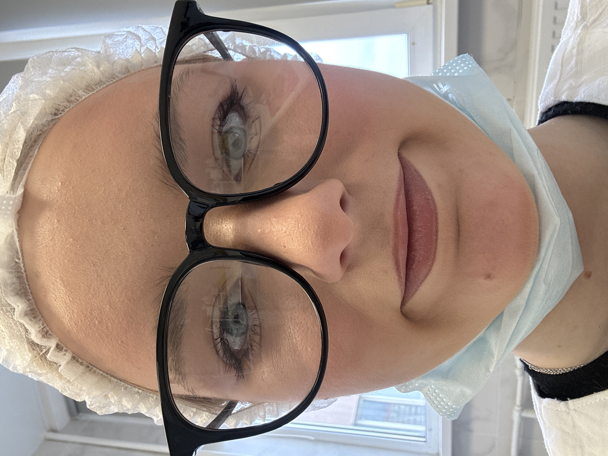 A close-up, head-on photograph captures the face of a young woman wearing a white surgical cap and black-rimmed glasses. Her light blue eyes are visible through the lenses, framed by dark eyelashes and light brown eyebrows. A light blue disposable face mask is partially covering her chin and mouth, which is slightly pursed. She appears to be wearing a white medical garment. The background consists of a window with blinds and a radiator, suggesting an indoor setting. No specific location within the city of Unknown, Russia is discernible from the image.